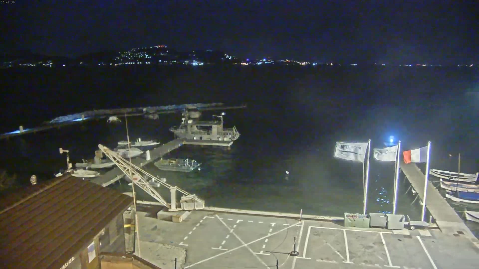 A nighttime view of a harbor with several boats docked, a crane, and a parking lot, under a clear, dark sky.