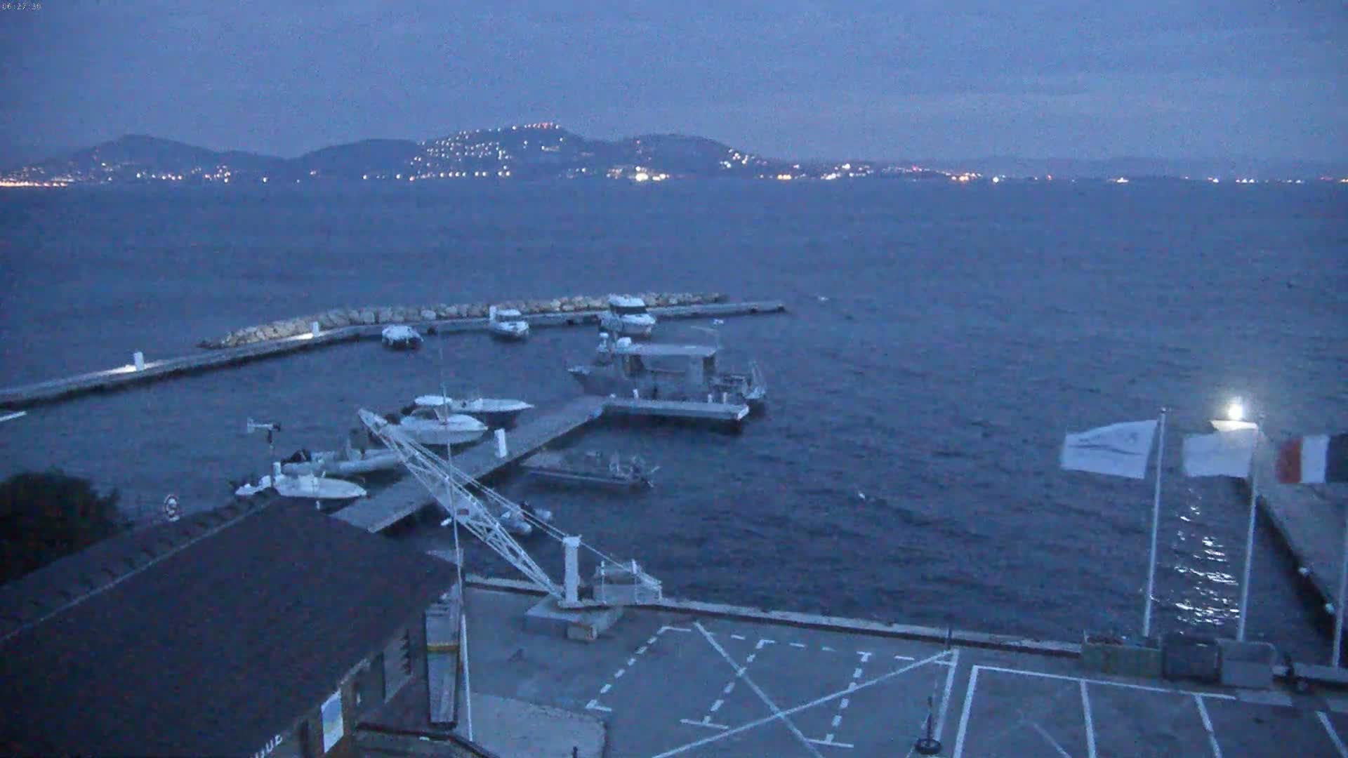 Several boats are docked at a marina at dusk, with a distant, illuminated coastline visible across the dark water.