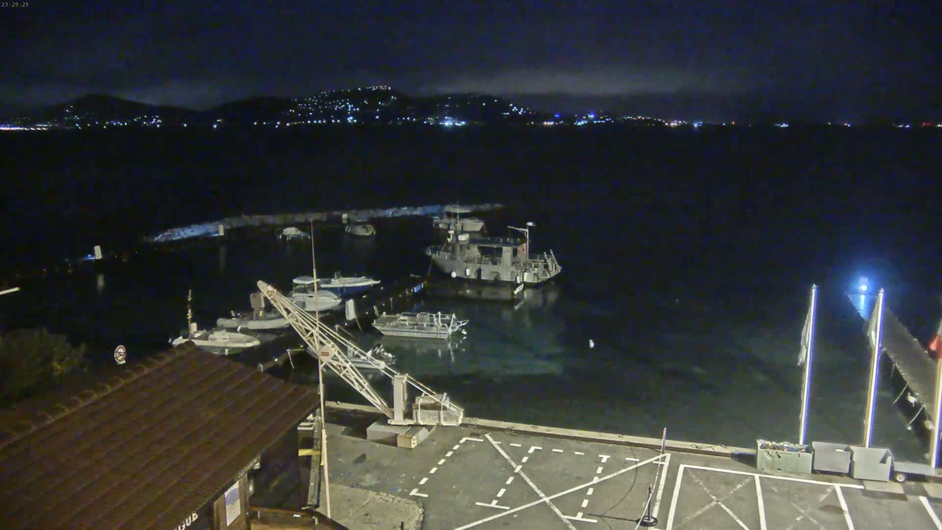 At night, several boats are moored in a calm harbor, with a city's lights visible across the water.
