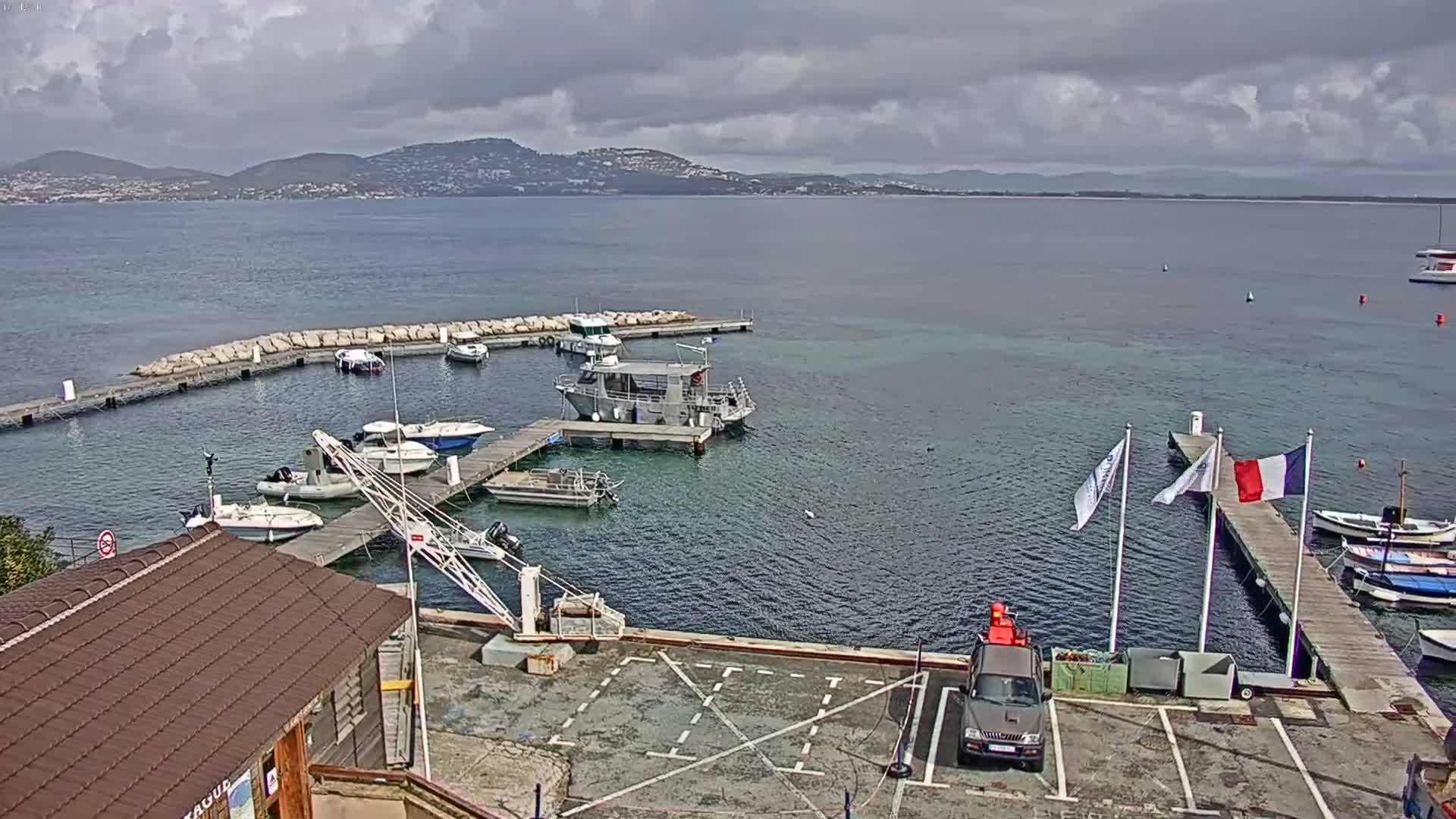 A small marina with several boats docked, a crane, and a building, overlooks a calm bay under a partly cloudy sky with distant hills visible in the background.