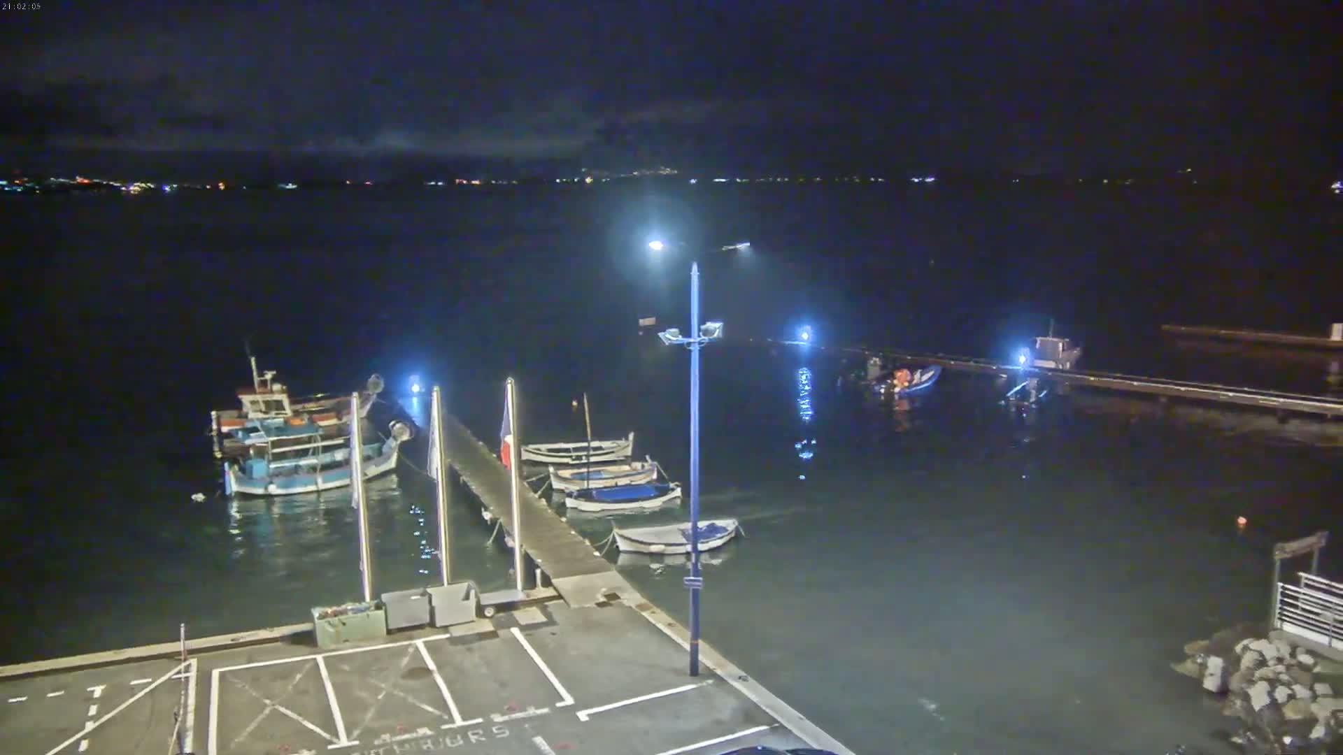 At night, several small boats are moored at a pier next to a dark body of water under a mostly clear sky.
