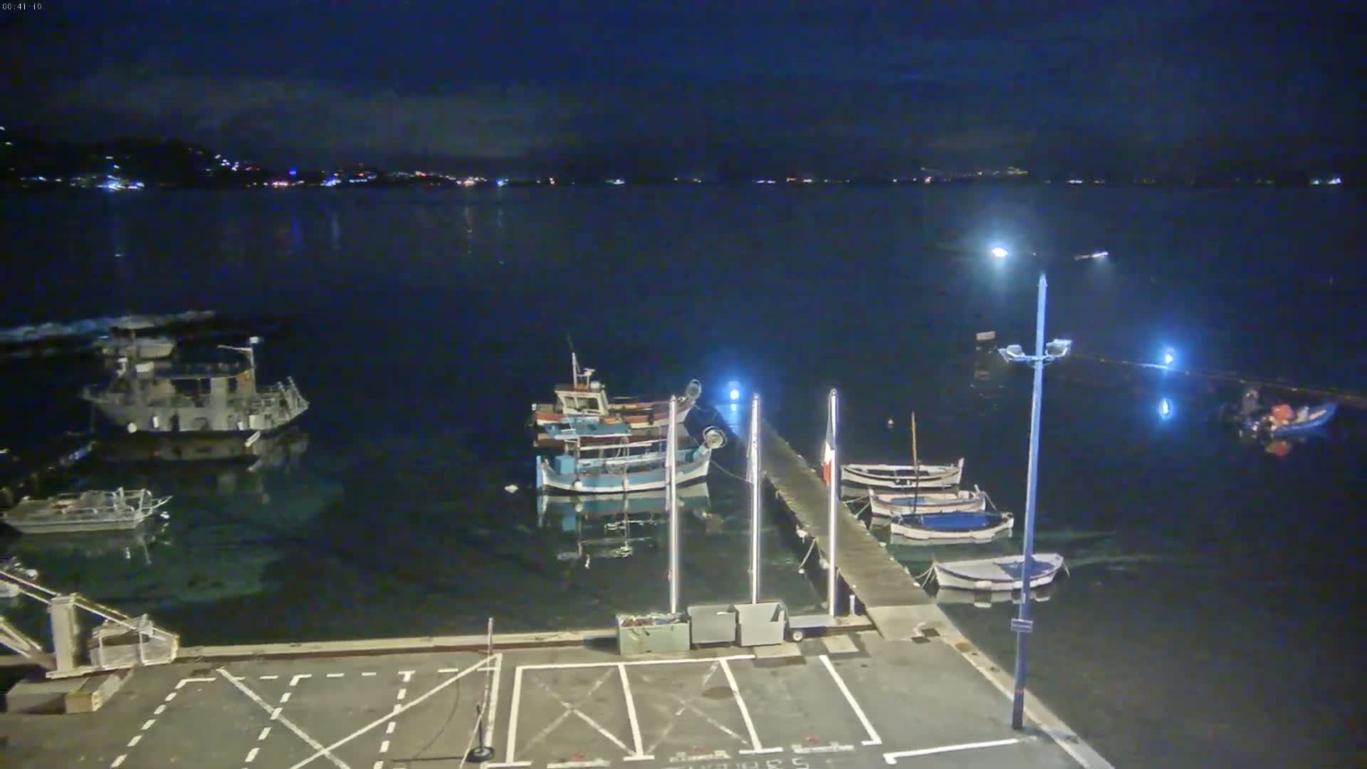 Several boats are moored at a pier at night, under a dark, clear sky, across from a dimly lit shoreline.