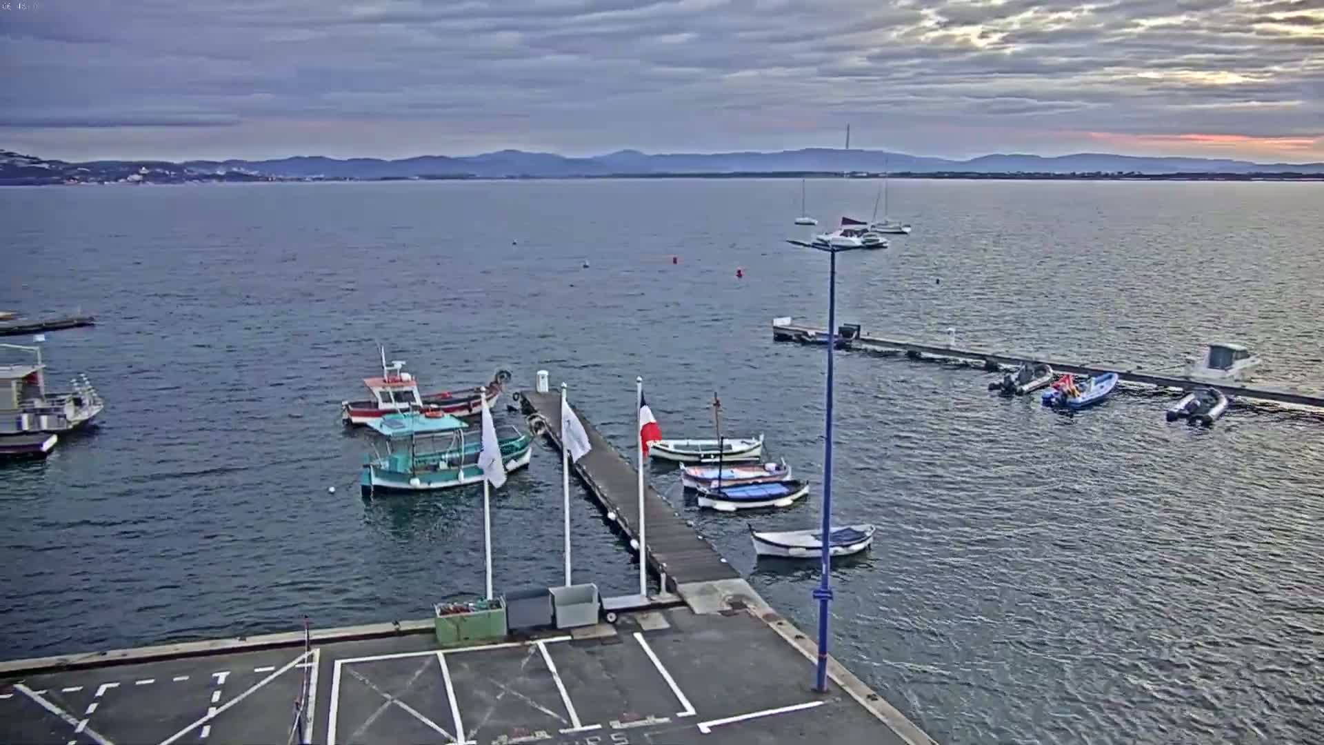 Several small boats are moored at a dock on calm water under a cloudy sky, with distant mountains visible on the horizon.