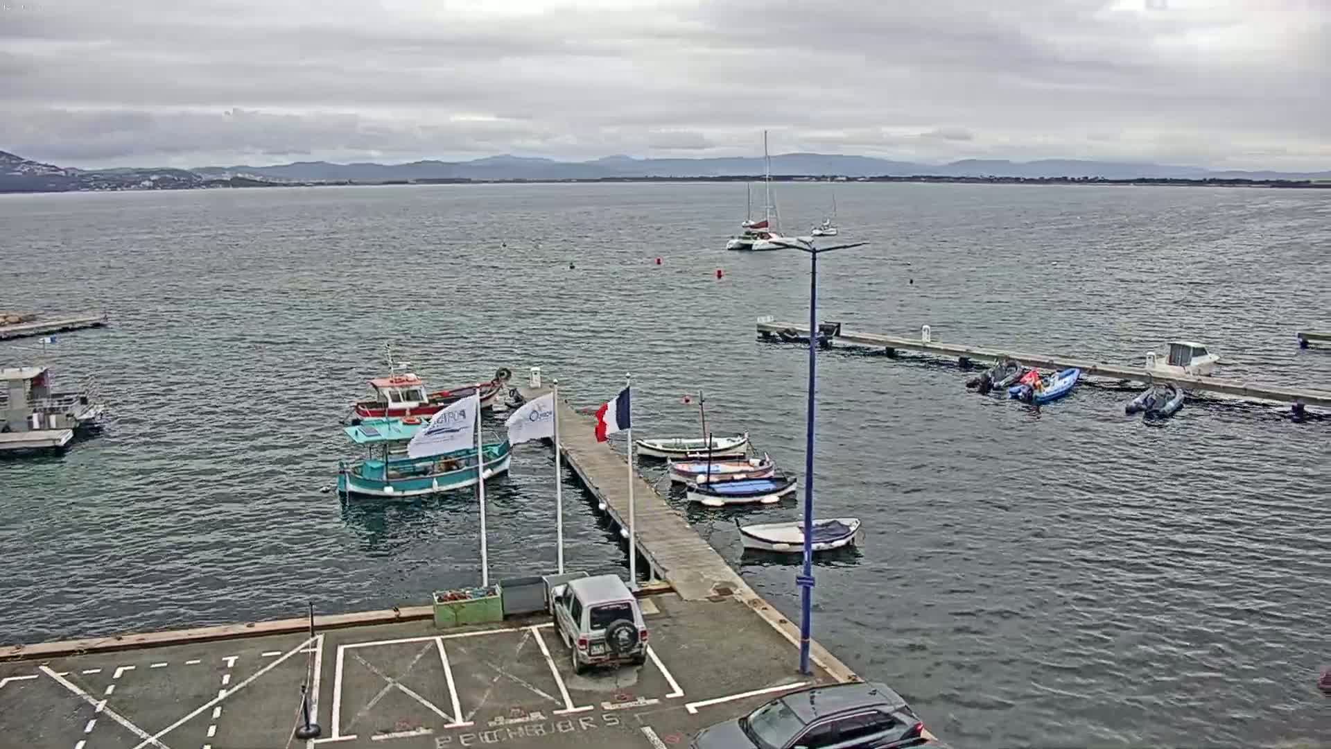 Several small boats are moored at a dock in calm water under an overcast sky, with a larger body of water and distant hills visible in the background.