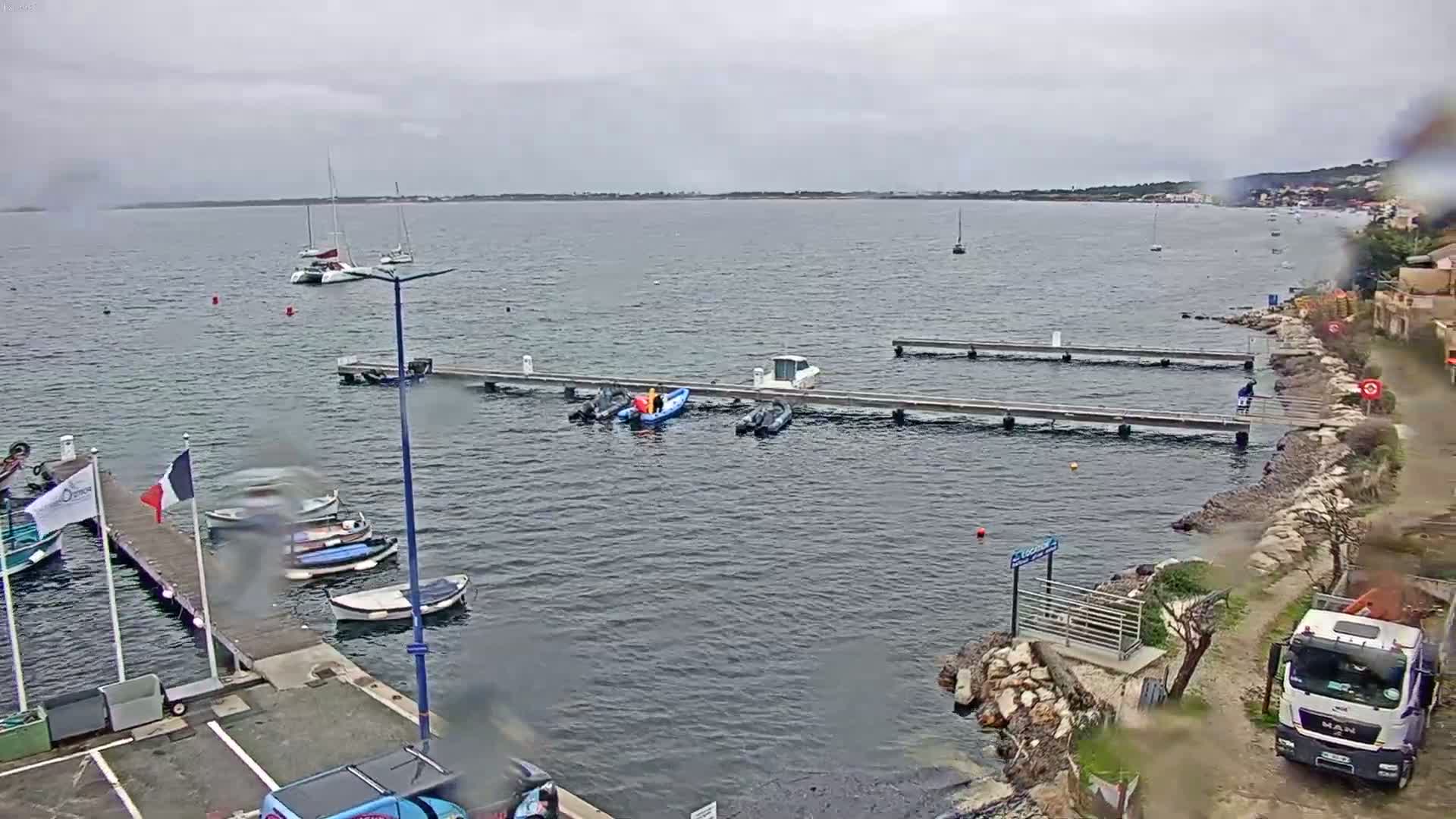 A small harbor with several boats moored at docks, a few people working on smaller boats, and a truck parked on the shore under an overcast sky.