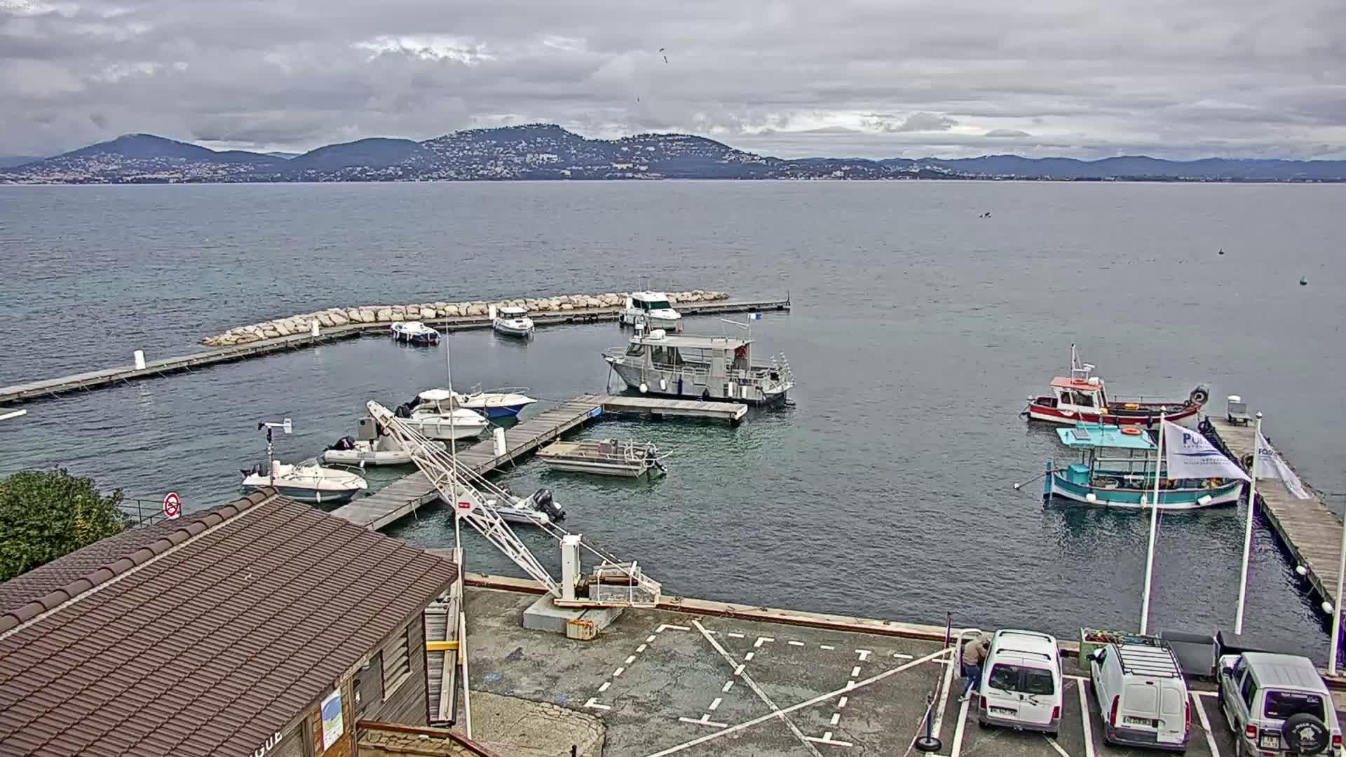 A small harbor filled with boats is nestled next to a building under a cloudy sky, with mountains visible in the distance across calm water.