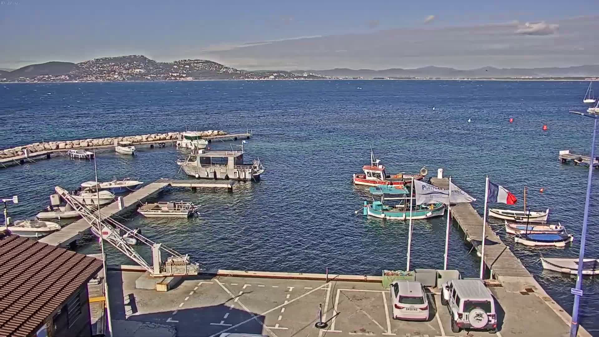 A small harbor with numerous boats docked at piers, overlooking a calm body of water and distant hills under a clear sky.