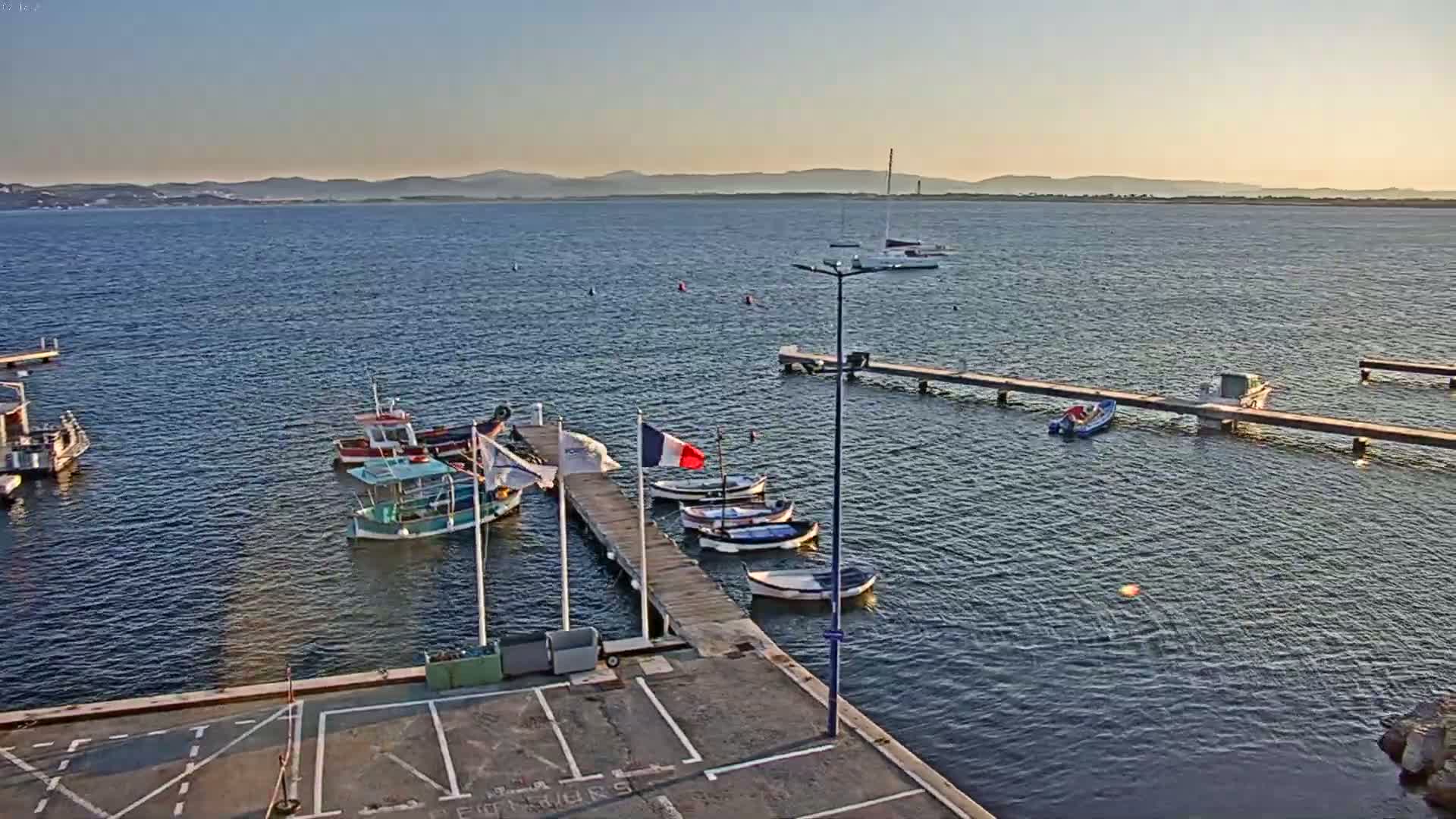 Several small boats are moored at a pier on calm water under a clear sky, with a distant mountain range visible on the horizon.