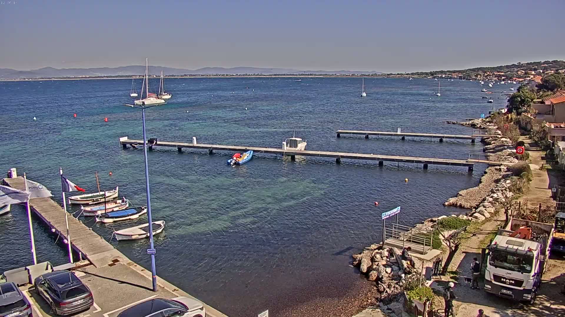 A sunny day at a harbor shows several small boats moored at docks, with a larger body of water extending to a distant shoreline.