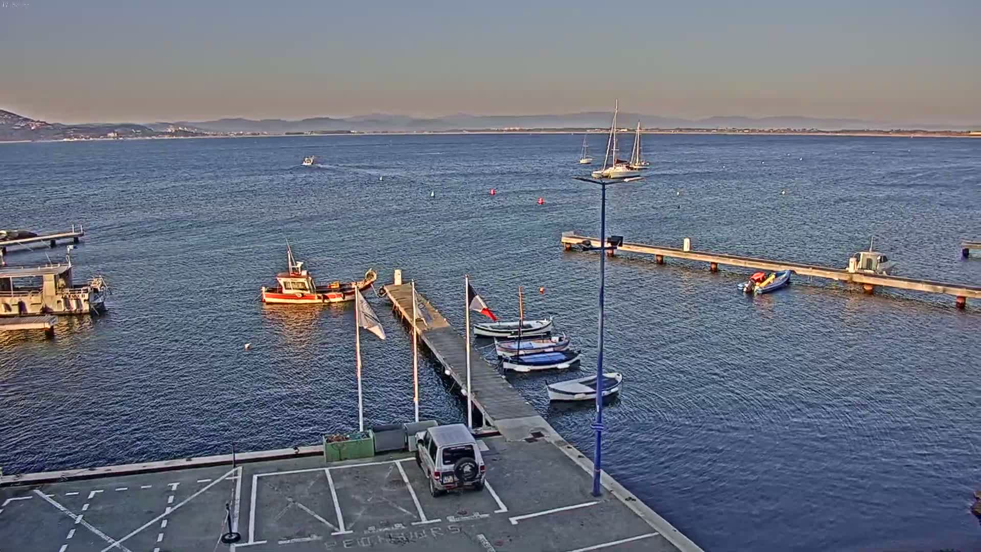 A calm, sunny day shows a small harbor with several boats docked at piers, a few sailboats on the water, and distant mountains under a clear sky.