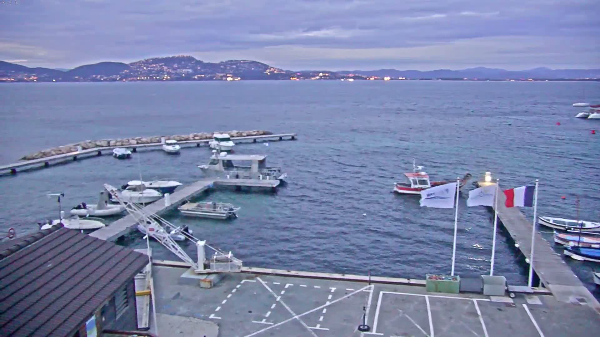 A small marina with several boats docked, under a cloudy, twilight sky, overlooks a calm body of water with distant hills and lights visible on the horizon.