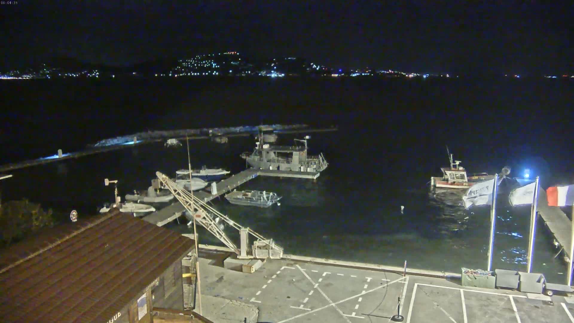 A nighttime view of a harbor with several boats docked, a crane, and a small building, under calm, dark conditions.