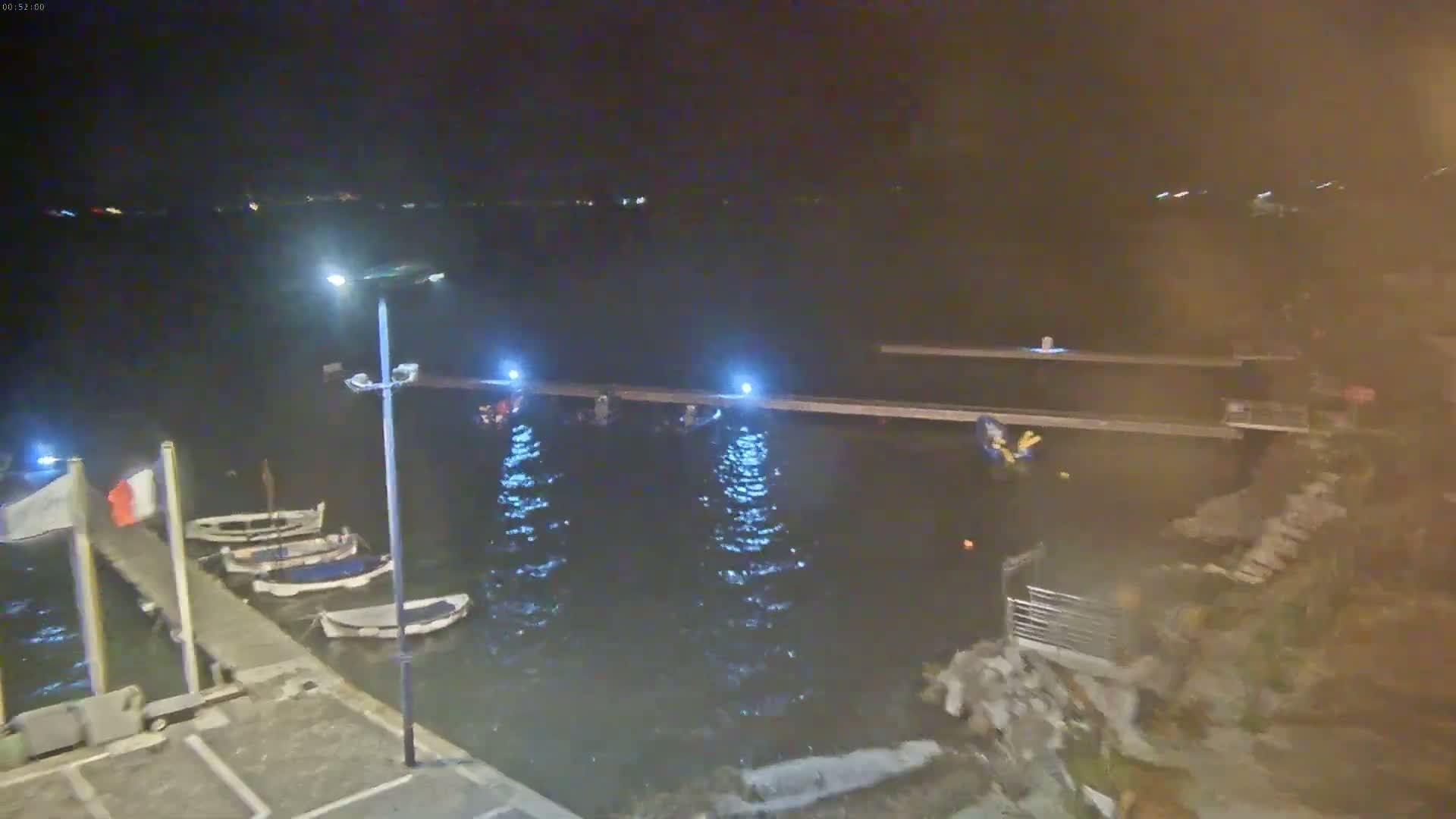 Several small boats are moored at a dock at night under a dark sky, with gently rippling water illuminated by lights along the dock.