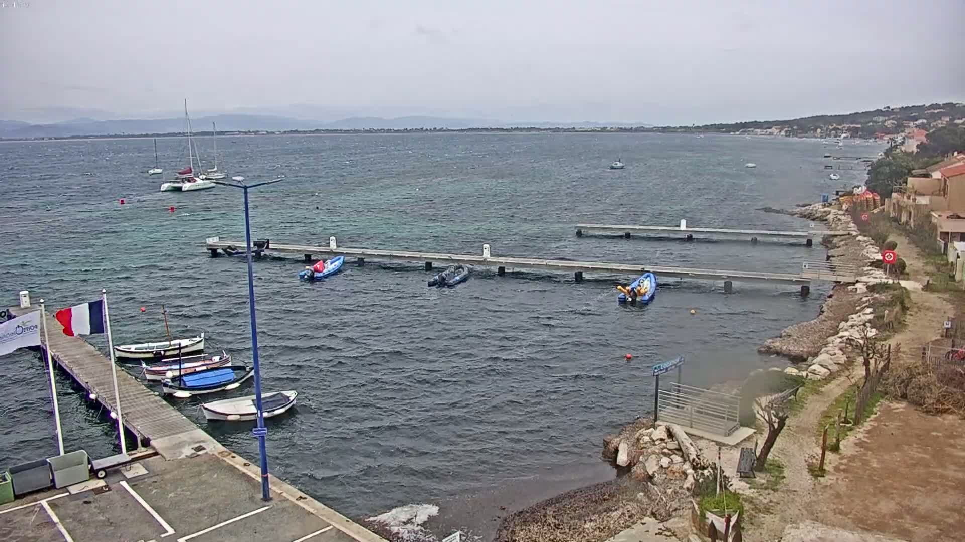 A partly cloudy day shows several small boats moored at a pier next to a calm, choppy body of water, with additional boats and sailboats visible further out.