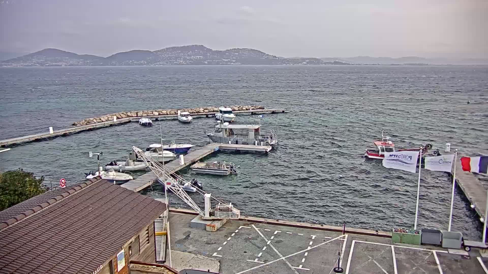 A small marina with numerous boats docked at piers, under a cloudy sky, with a mountainous coastline visible in the distance across choppy water.