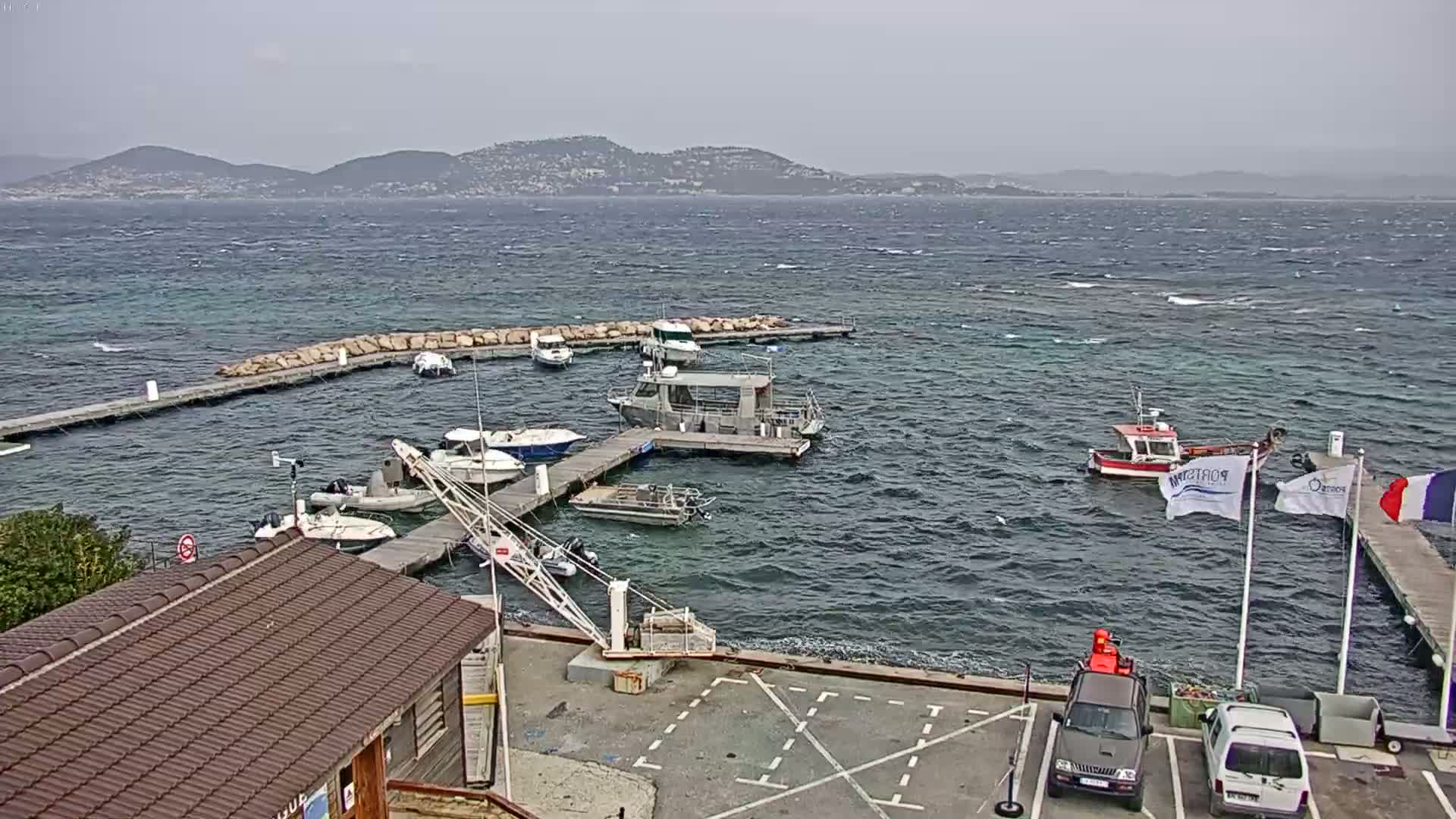 A small harbor with several boats moored at docks, under a cloudy sky with a windy sea showing small waves, and distant mountains in the background.