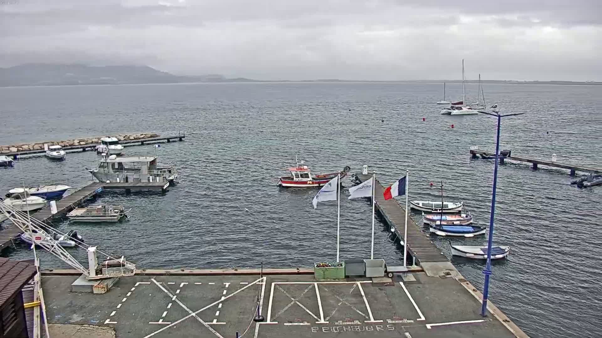 A small marina with numerous boats, including sailboats and small motorboats, is shown on a cloudy day with calm water and distant mountains visible on the horizon.