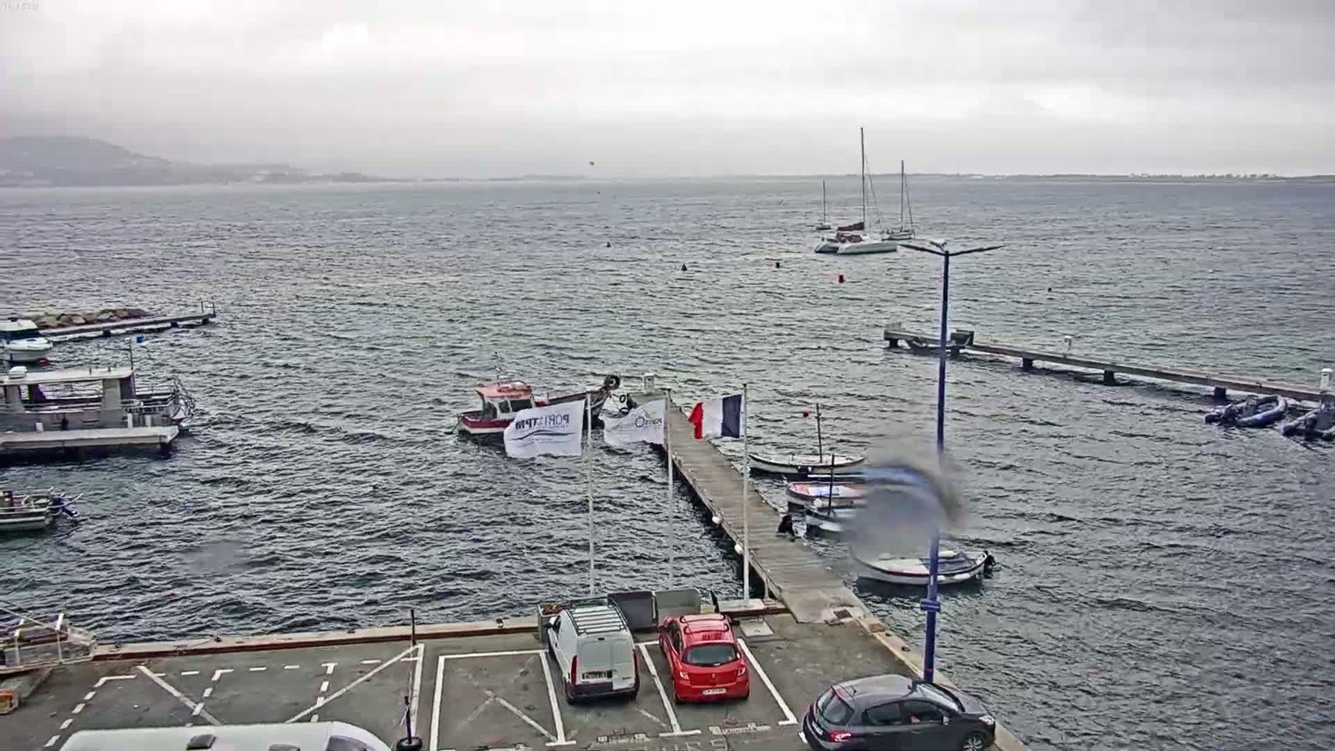 A harbor scene on an overcast day shows several boats moored at docks, with additional boats and cars in a parking lot.