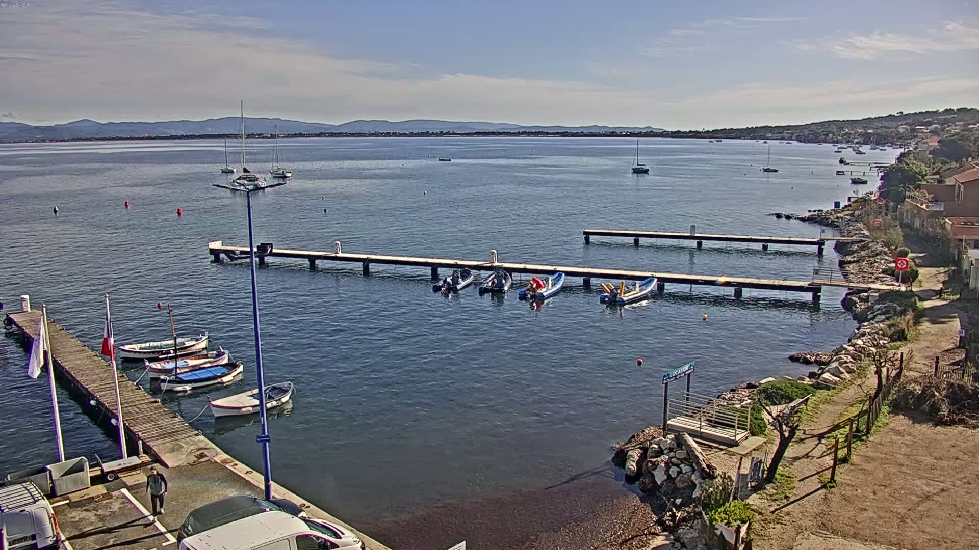 A calm bay with several boats moored at wooden piers, under a mostly sunny sky with some clouds.