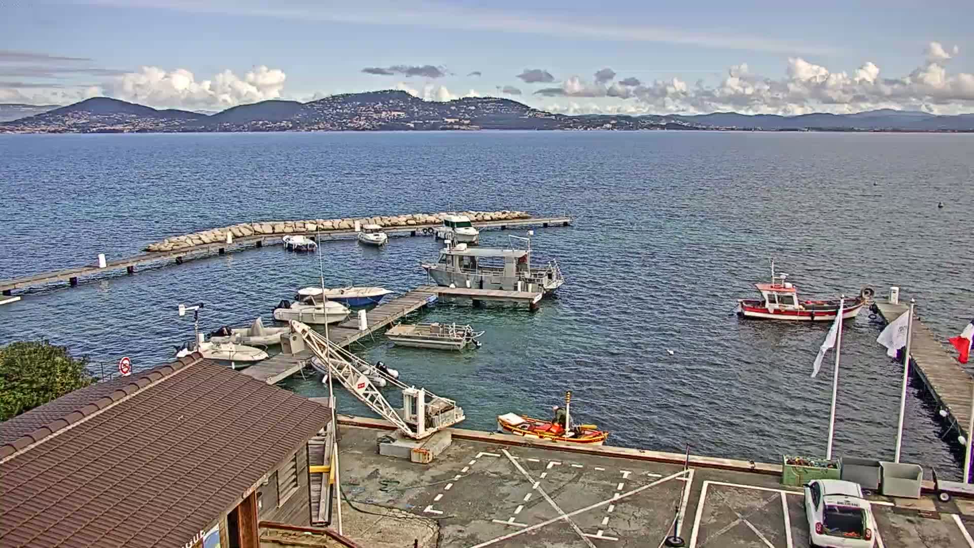 A small harbor with numerous boats docked at piers, a crane, and a building, is situated on calm water under a partly cloudy sky with distant mountains visible.