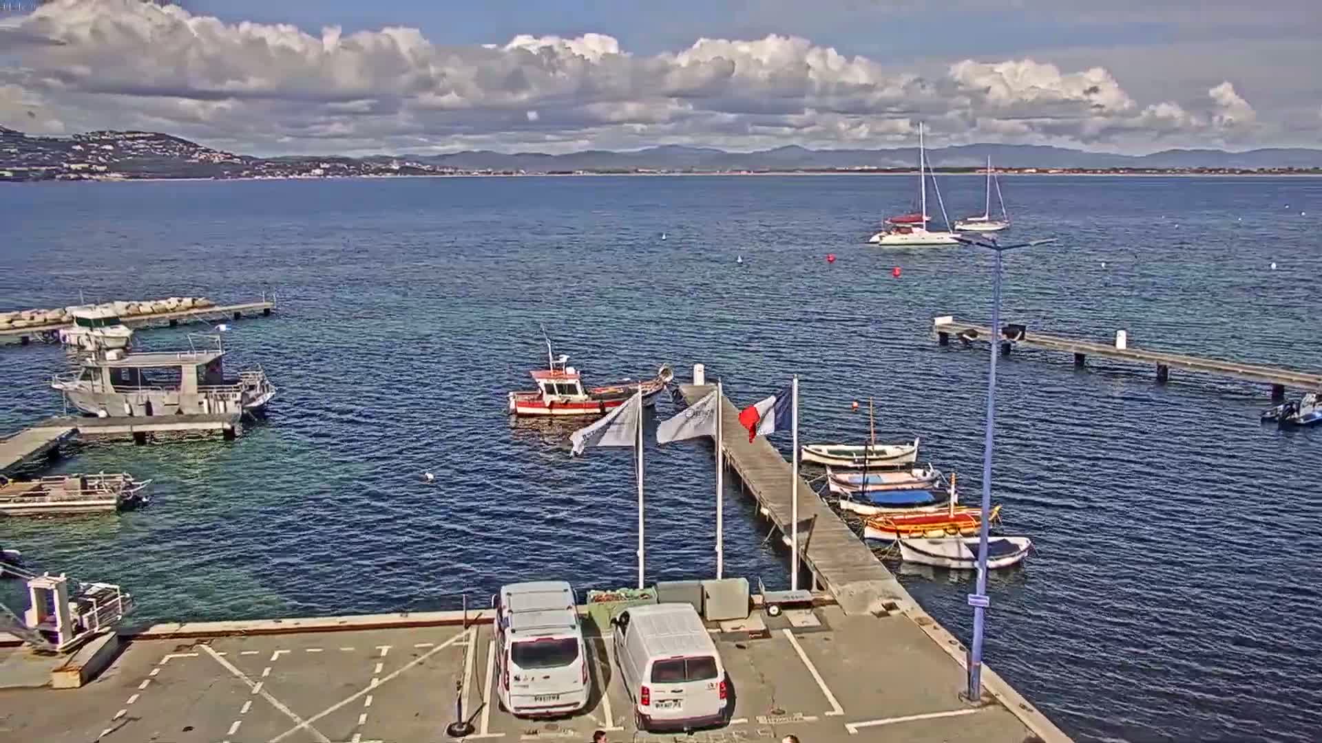 A partly cloudy day shows a marina with several boats moored at docks, two sailboats in the distance, and two vans parked in a lot beside the water.