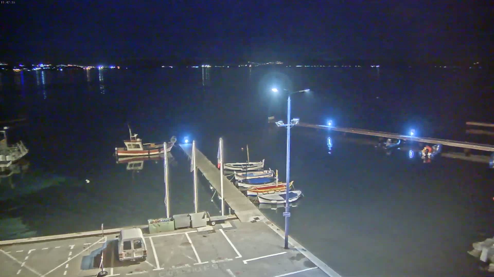 At night, several small boats are moored at a pier next to a calm body of water, with lights illuminating the scene and a distant shoreline visible.