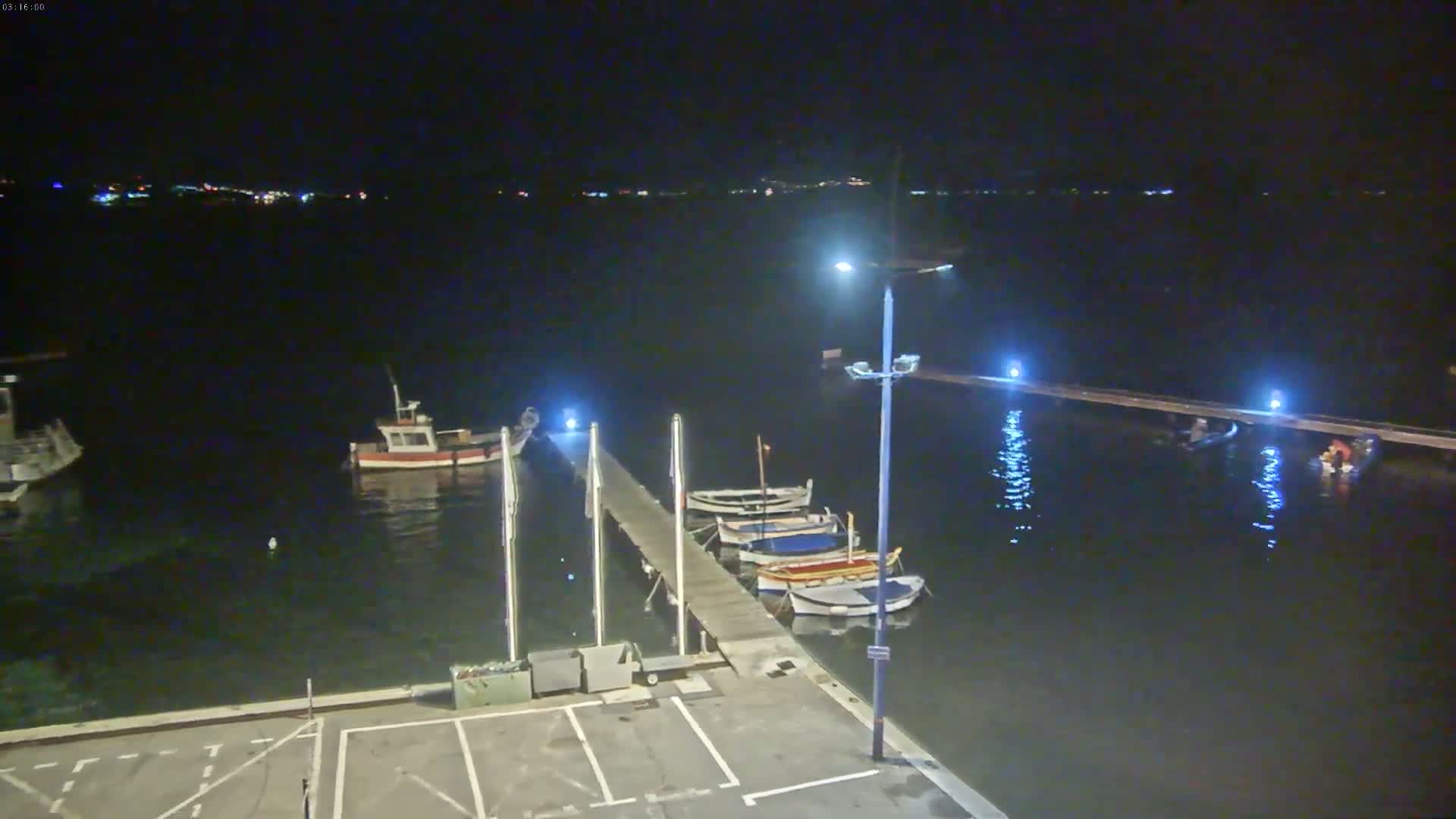 At night, several small boats are moored at a dock in calm water, with a few additional boats visible further out in the bay under a dark sky.