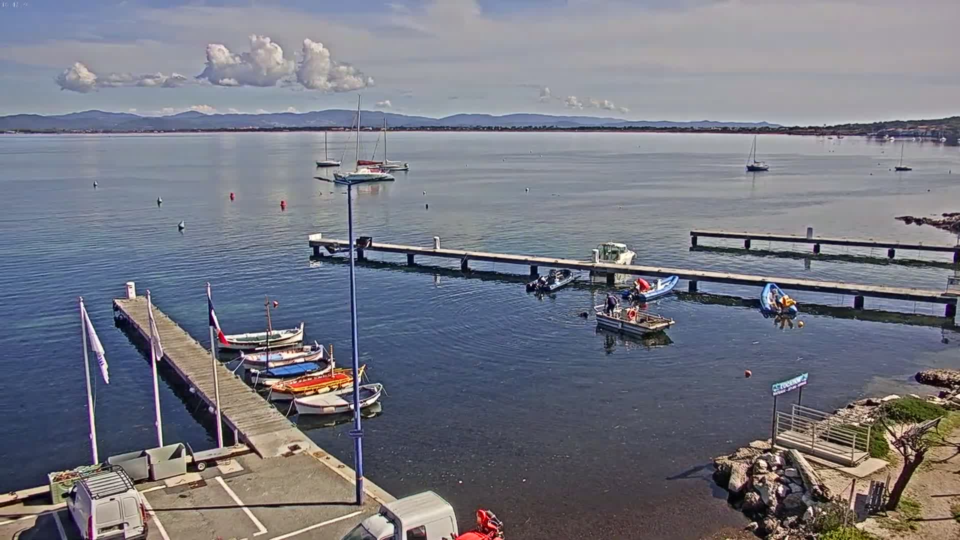 A calm, sunny day shows a harbor with several boats moored at docks, some people working on smaller boats in the water, and a distant mountain range visible on the horizon.