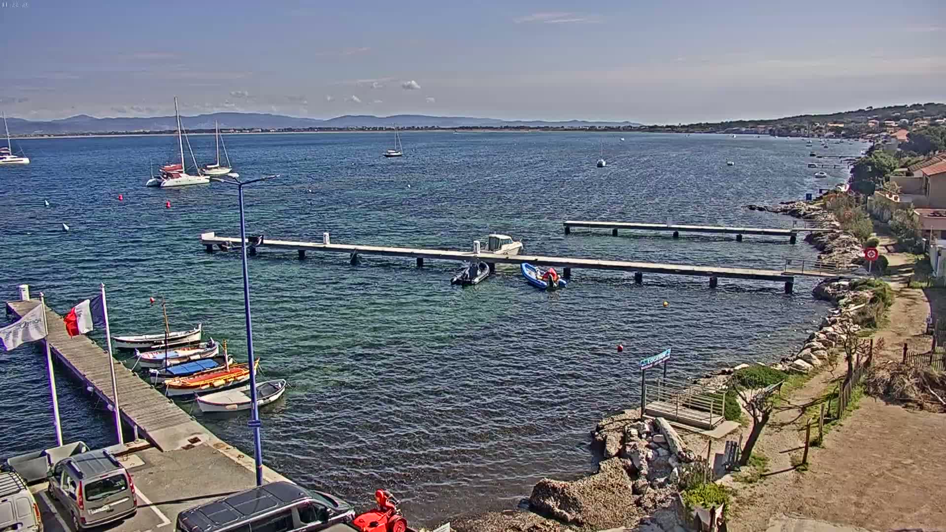 A calm, sunny day shows a marina with several boats docked at piers, extending into a bay with more boats and a distant coastline.