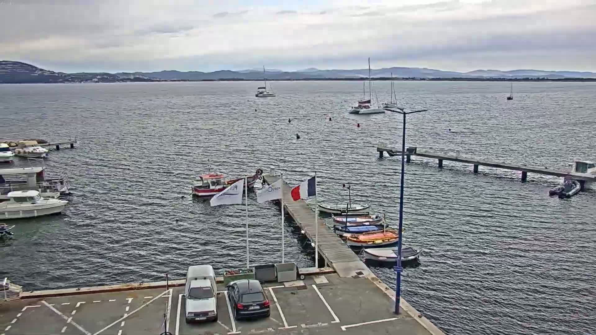 A calm, overcast day reveals a harbor scene with several boats docked at piers and others sailing on the water, with distant hills visible on the horizon.