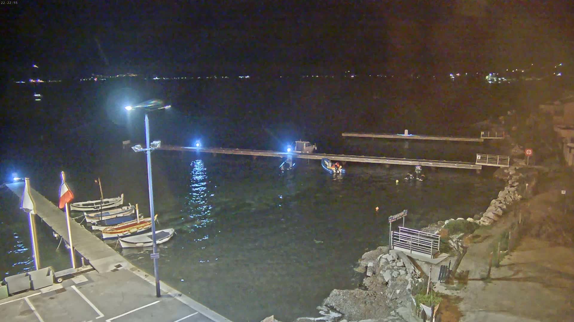 Several small boats are moored at a dock at night, with a few people visible near the water's edge under artificial light.