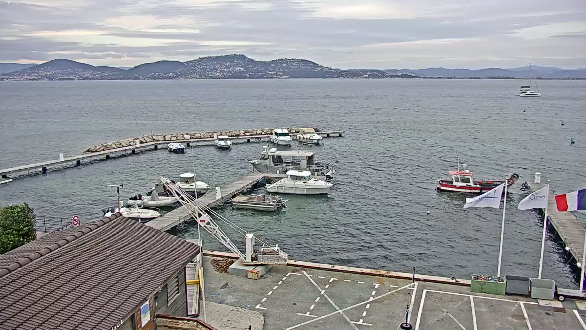 A small marina with numerous boats docked at piers, a small crane, and a building, overlooks a calm body of water with mountains in the distance under an overcast sky.
