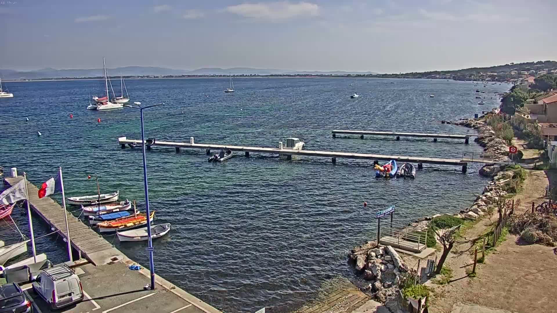 A sunny day at a harbor shows several boats moored at docks extending from a rocky shore, with additional boats and sailboats in the calm water beyond.