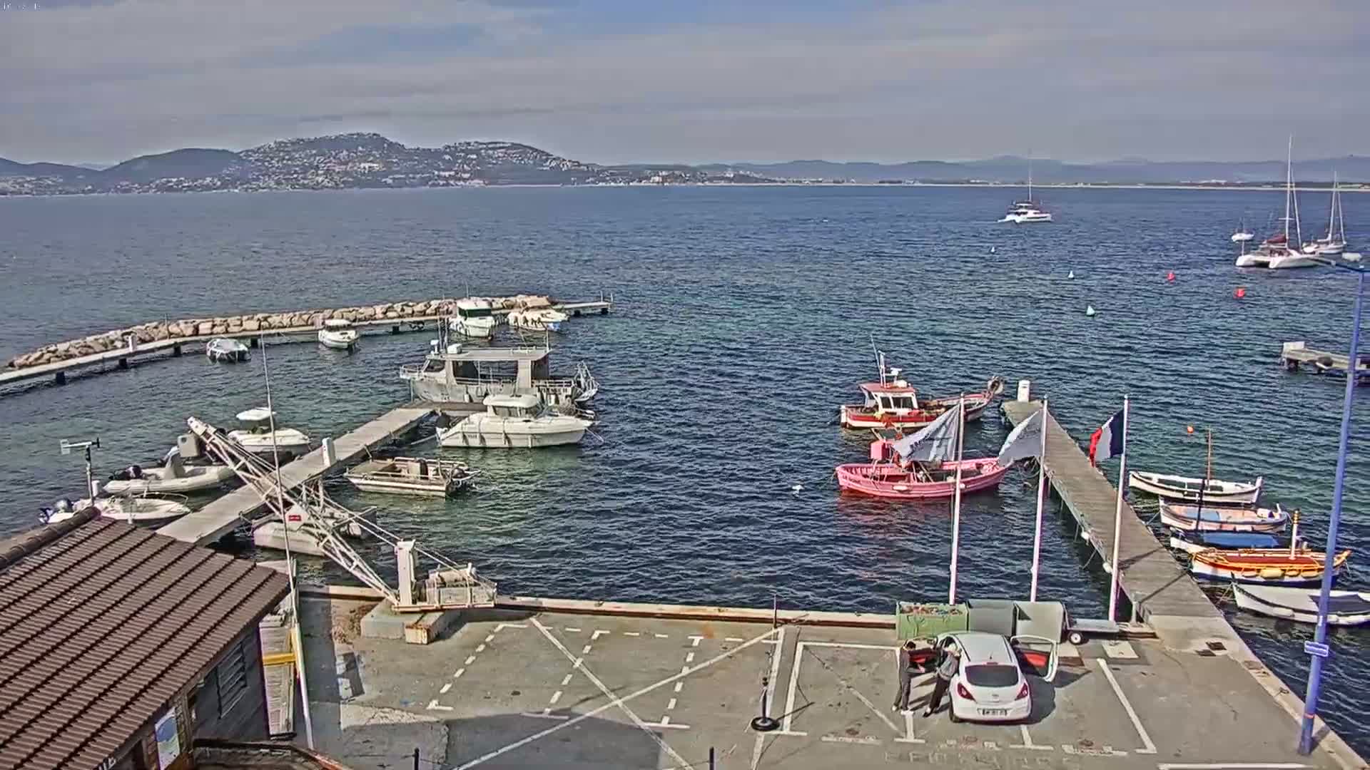 A small marina with numerous boats docked, a few cars in a parking lot, and a calm body of water with distant mountains under a partly cloudy sky.