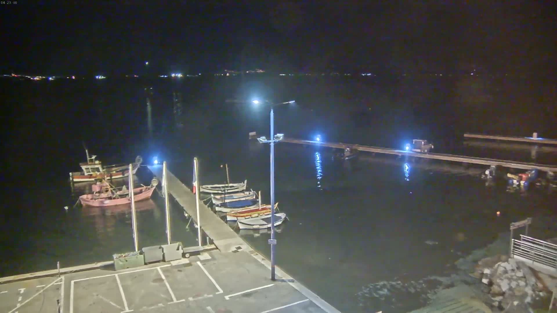 At night, several small boats are moored at a dock beside calm water, with a few more boats visible farther out in the water.