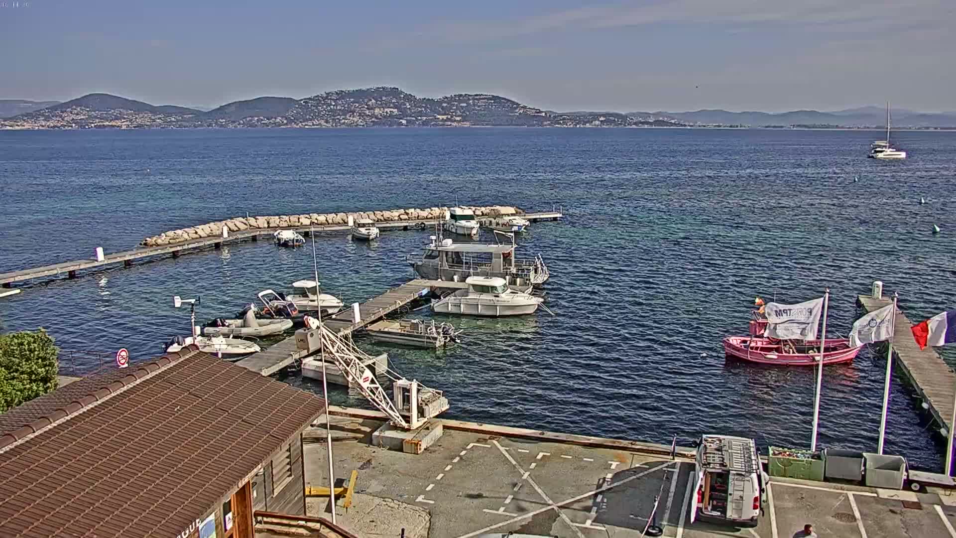 A small harbor filled with boats on a sunny day, with a larger body of water and distant mountains in the background.
