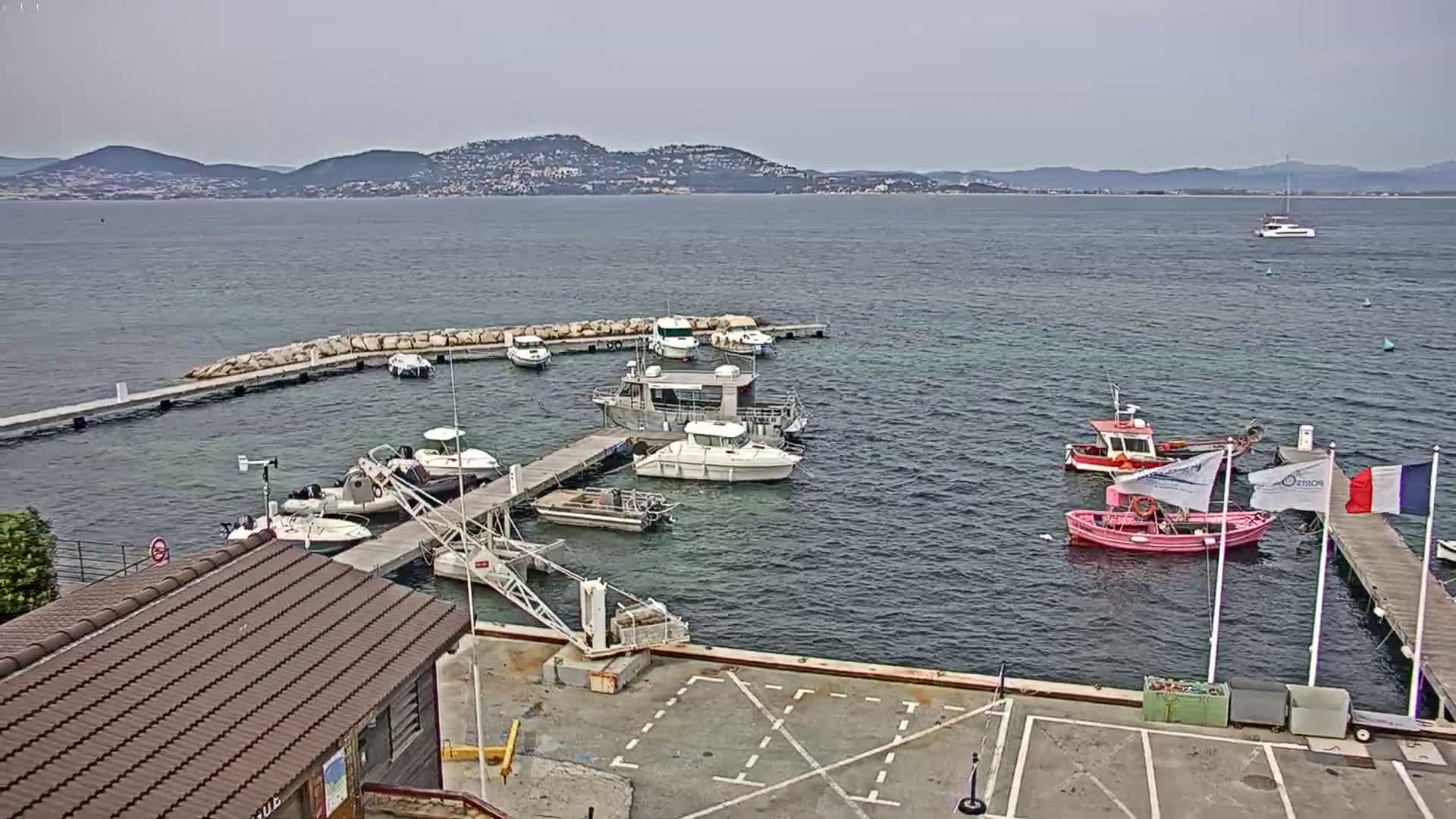 A small marina with numerous boats docked at piers, nestled in a calm bay under an overcast sky with distant mountains visible on the horizon.