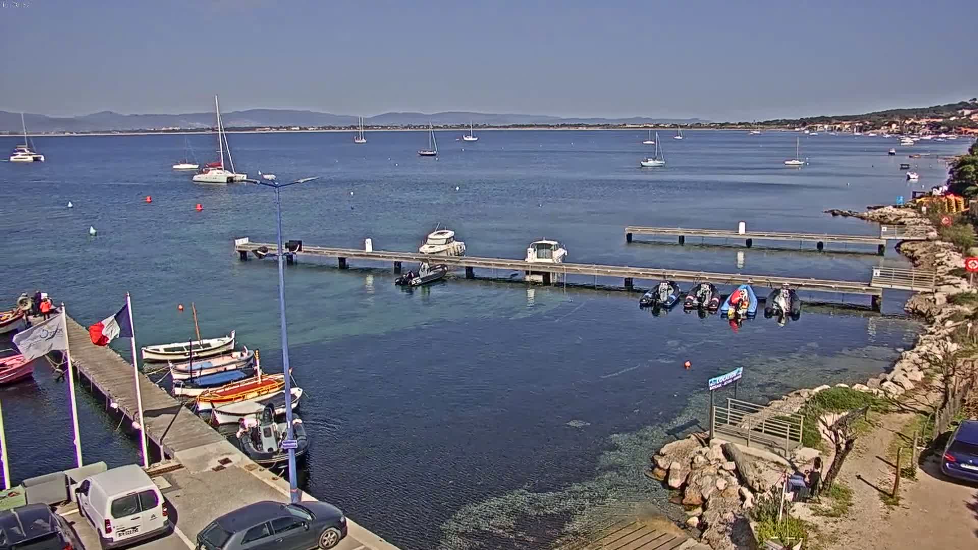 A harbor with numerous boats docked at piers, under a clear sunny sky, shows several other boats in the calm water beyond.