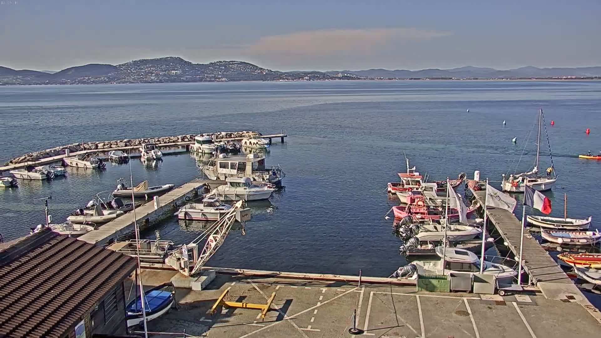 A harbor filled with numerous boats of varying sizes is nestled against a calm body of water under a clear, sunny sky, with mountains visible in the distance.