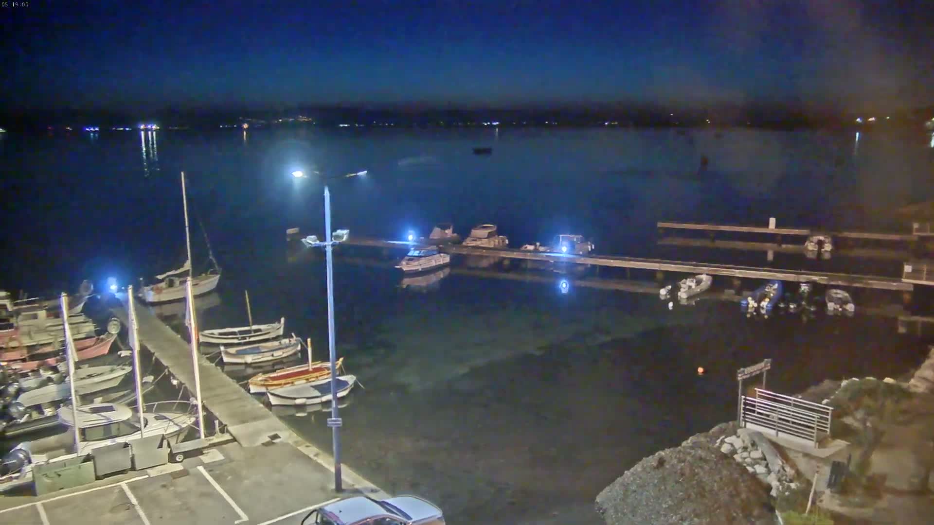 Under a dark night sky, numerous boats are moored at a dock in calm water, illuminated by several lights.