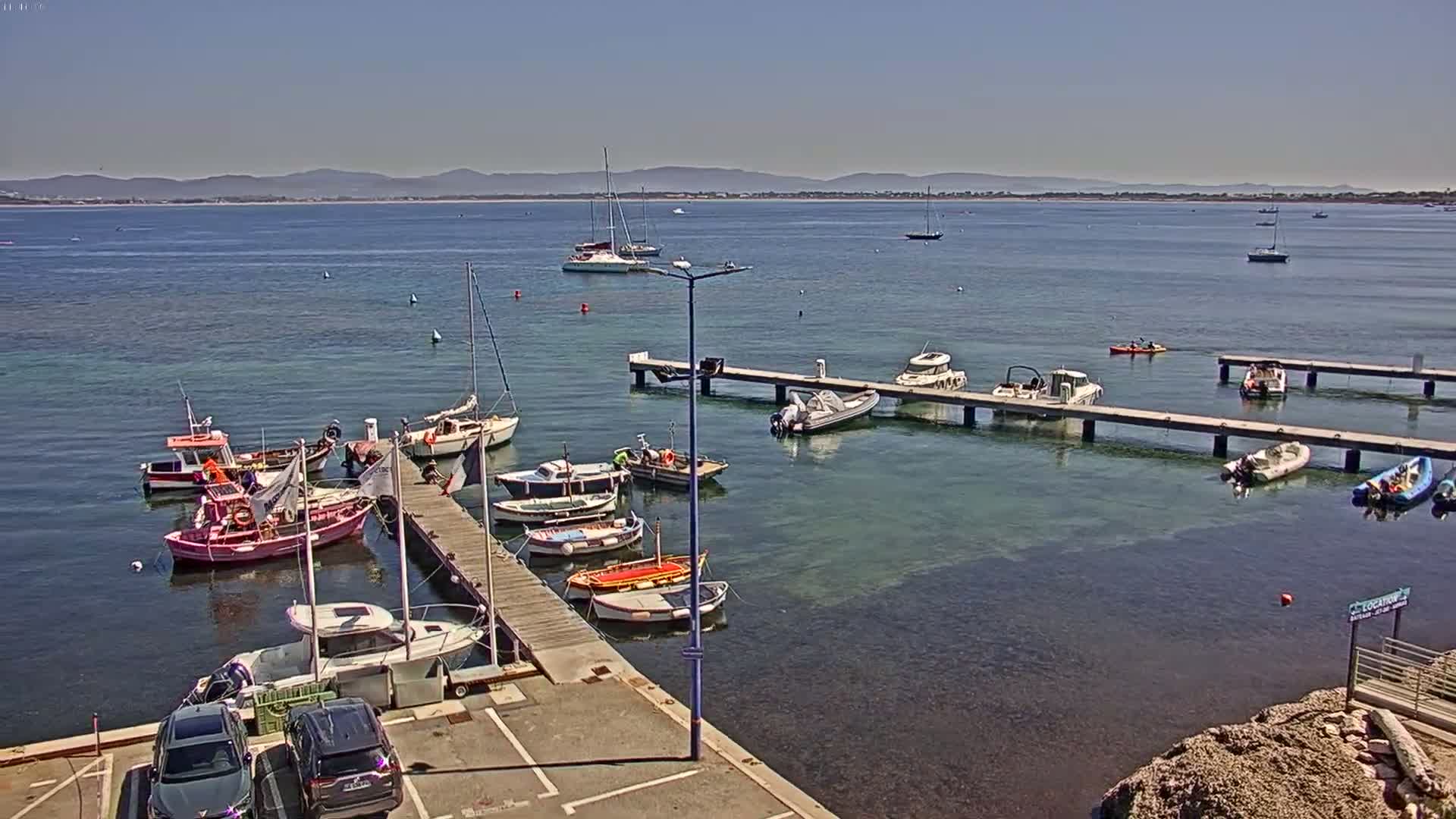 A sunny day shows a harbor with numerous boats moored at wooden docks, some people are visible near the boats, and a clear view of distant mountains across the calm water.