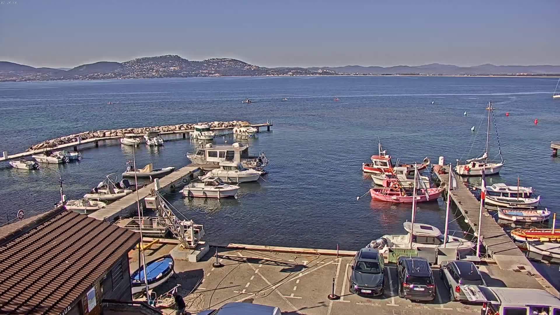 A small marina filled with various sized boats and cars parked in a lot next to it, all under a clear sunny sky, with a large body of water and distant hills in the background.