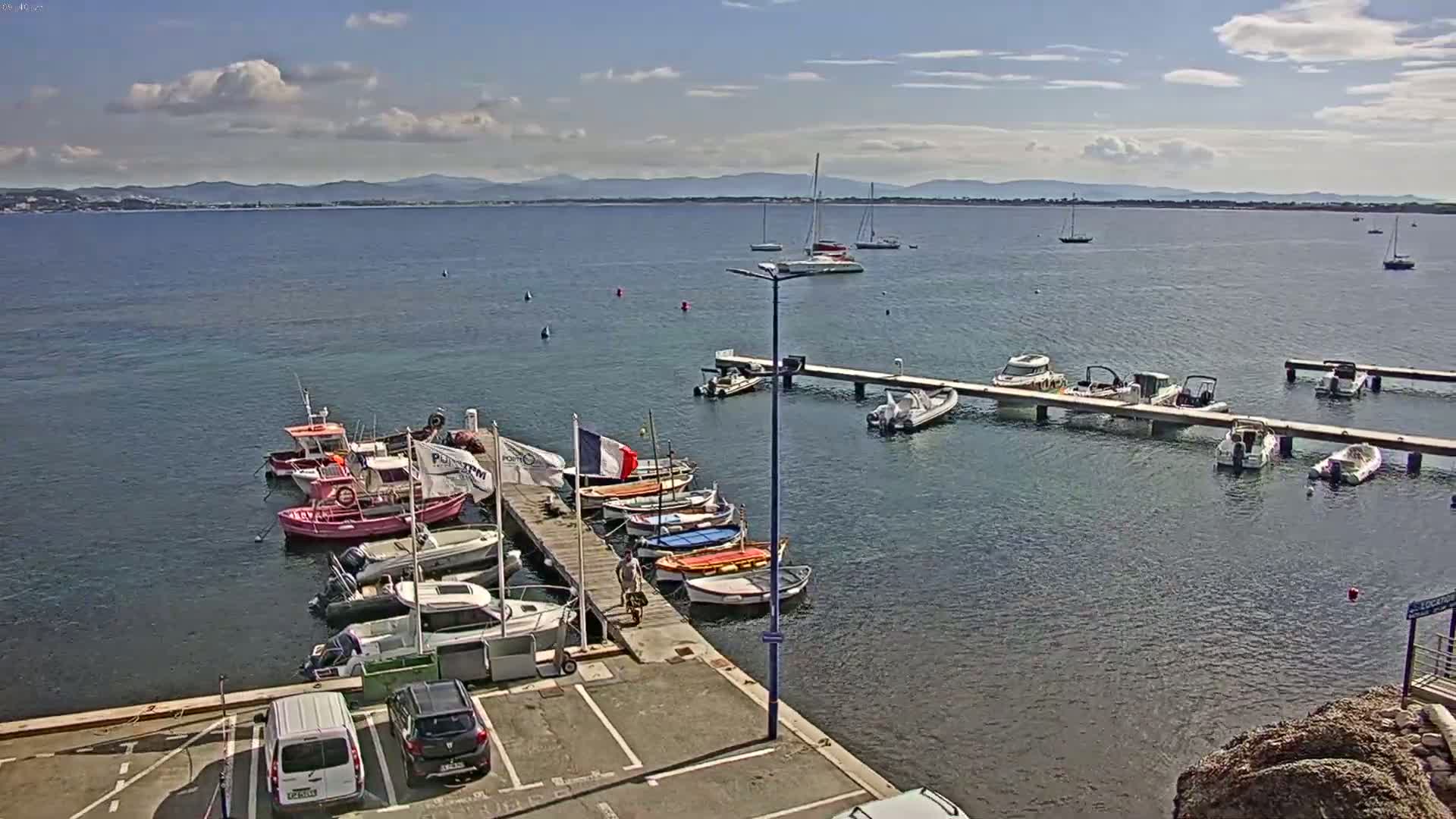 A partly cloudy day shows a marina with numerous small boats and yachts docked along piers, with a calm body of water extending to distant mountains.