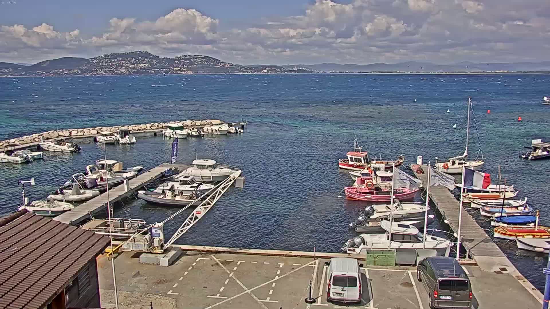 A marina filled with numerous boats of varying sizes is situated near a coastline under a partly cloudy sky, with a mountainous region visible in the distance.