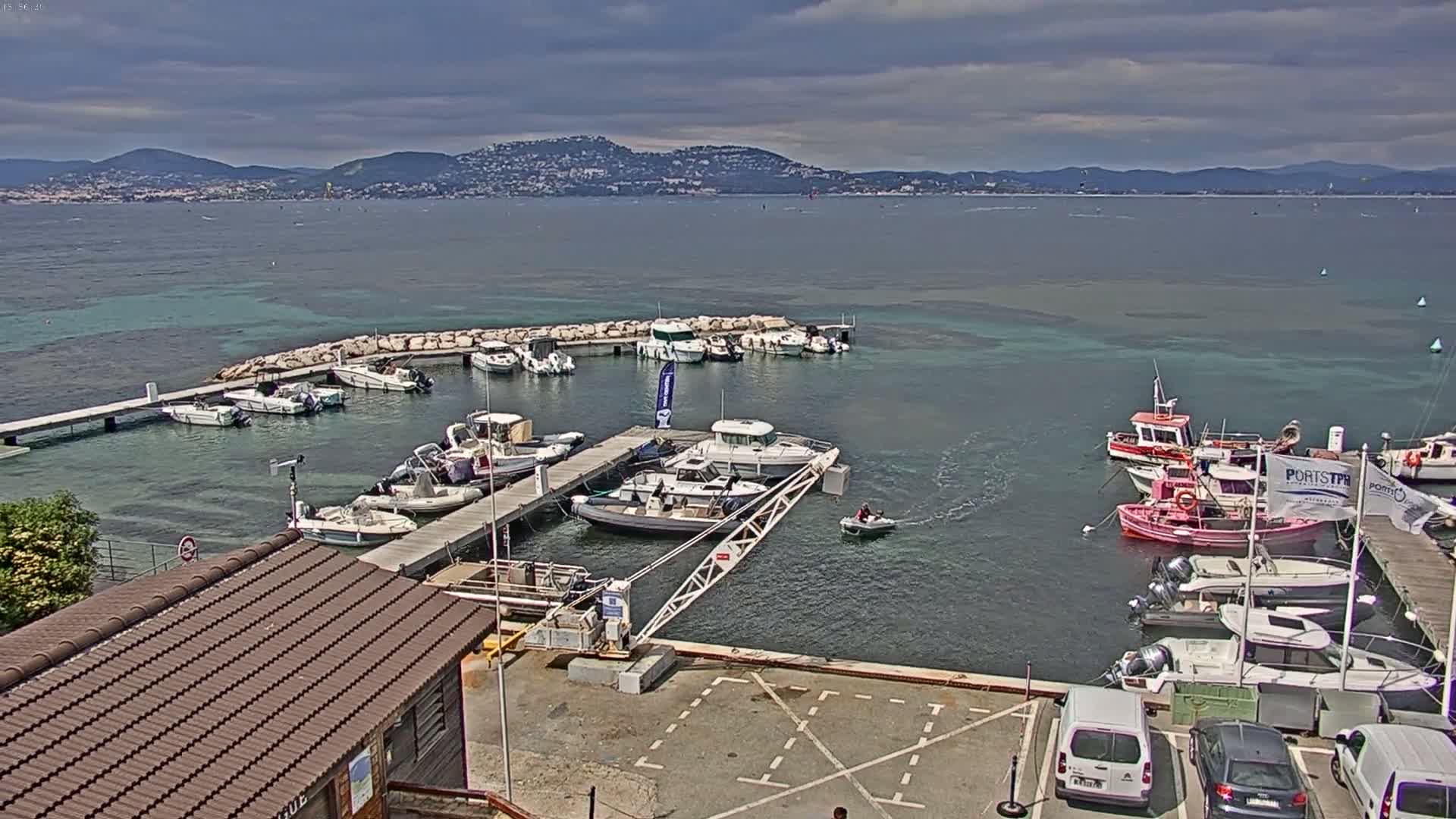 A small marina filled with various boats is situated near a shoreline, with a mountainous landscape visible in the distance under a partly cloudy sky.