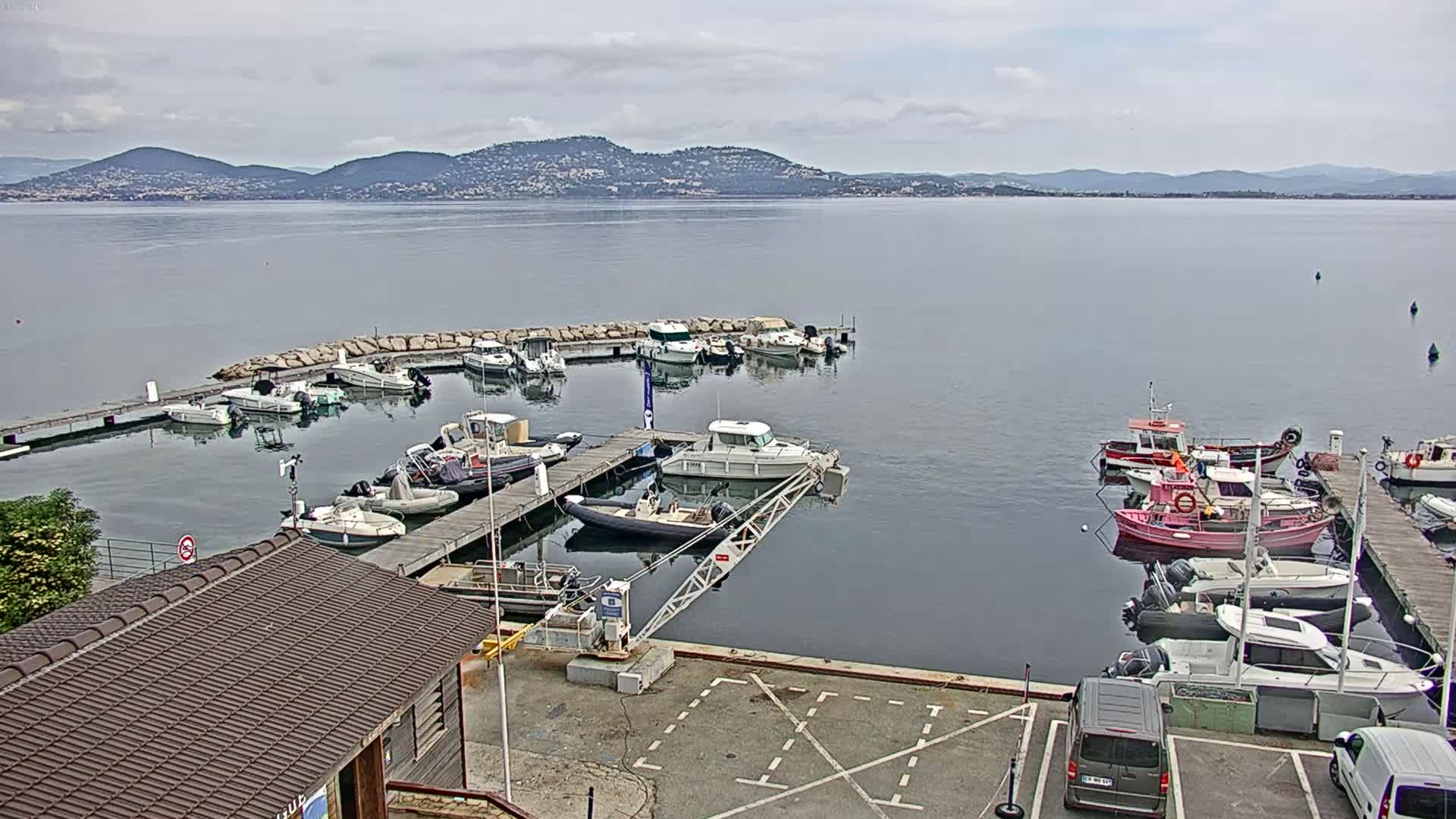 A calm body of water with numerous boats docked at a marina, mountains visible in the distance under a cloudy sky.