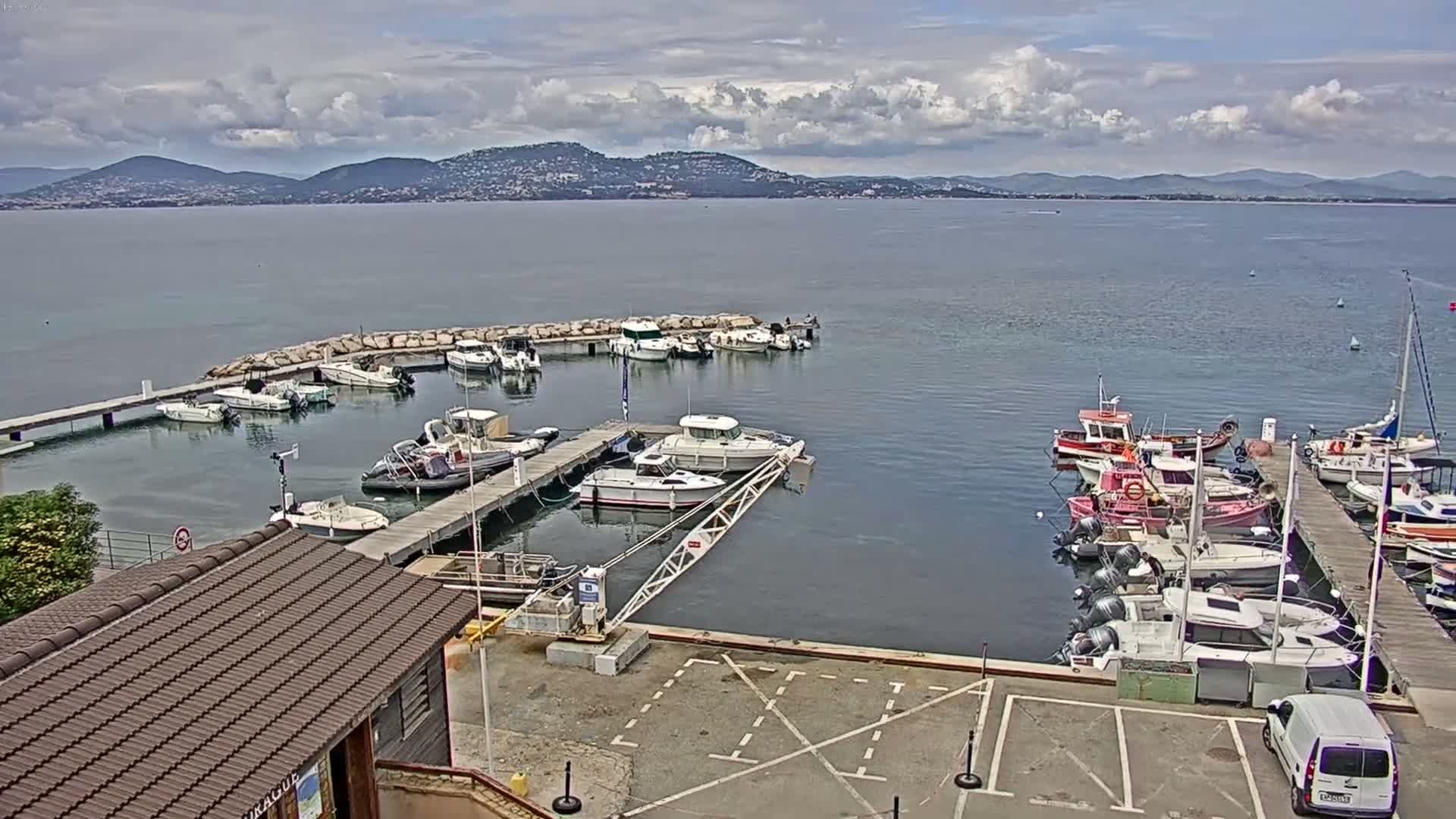 A marina filled with numerous boats of varying sizes is situated on calm water under a partly cloudy sky, with distant mountains visible in the background.