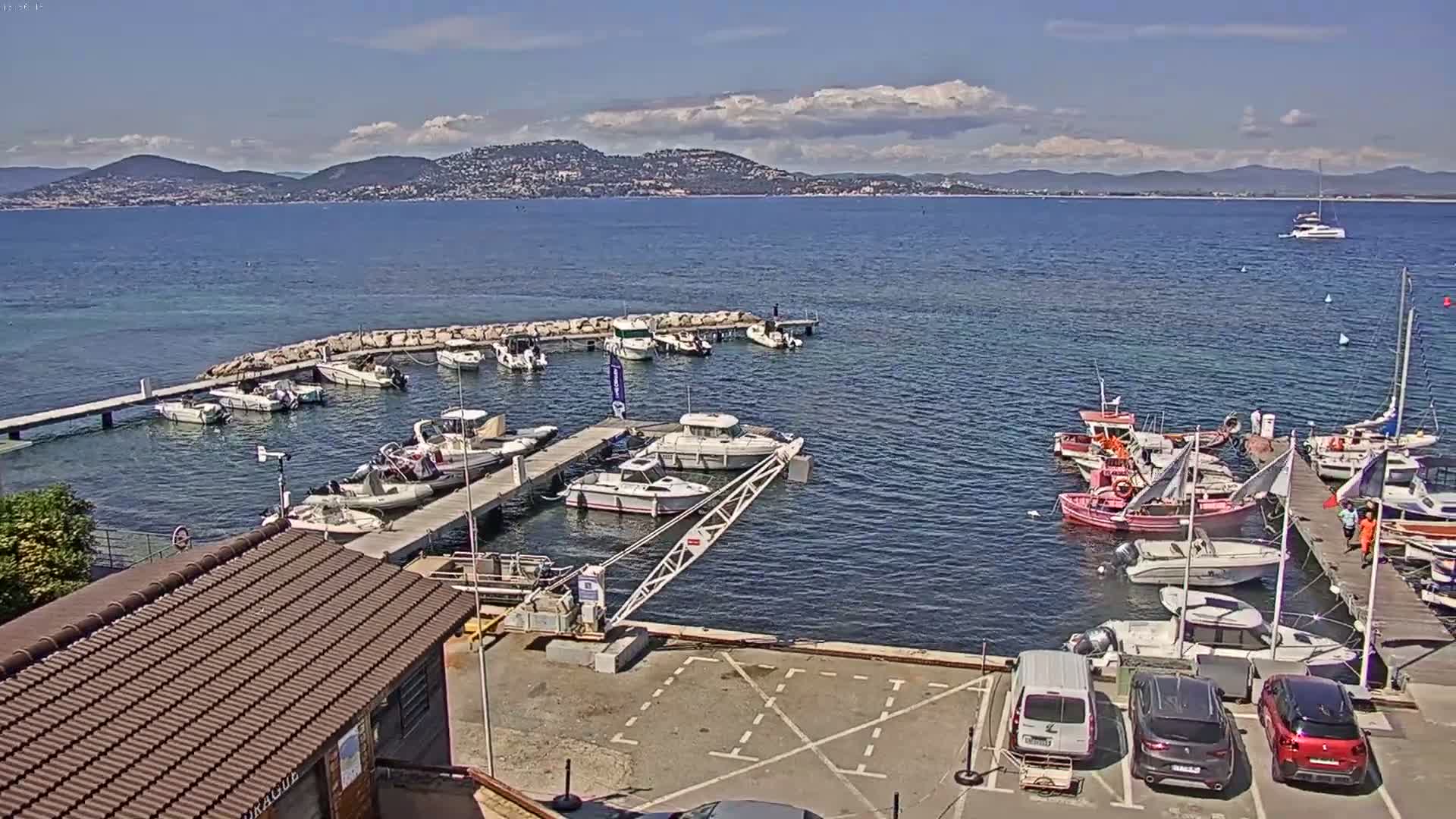 A marina filled with various boats, including sailboats and motorboats, is situated on calm blue water under a mostly sunny sky, with mountains visible in the distance.