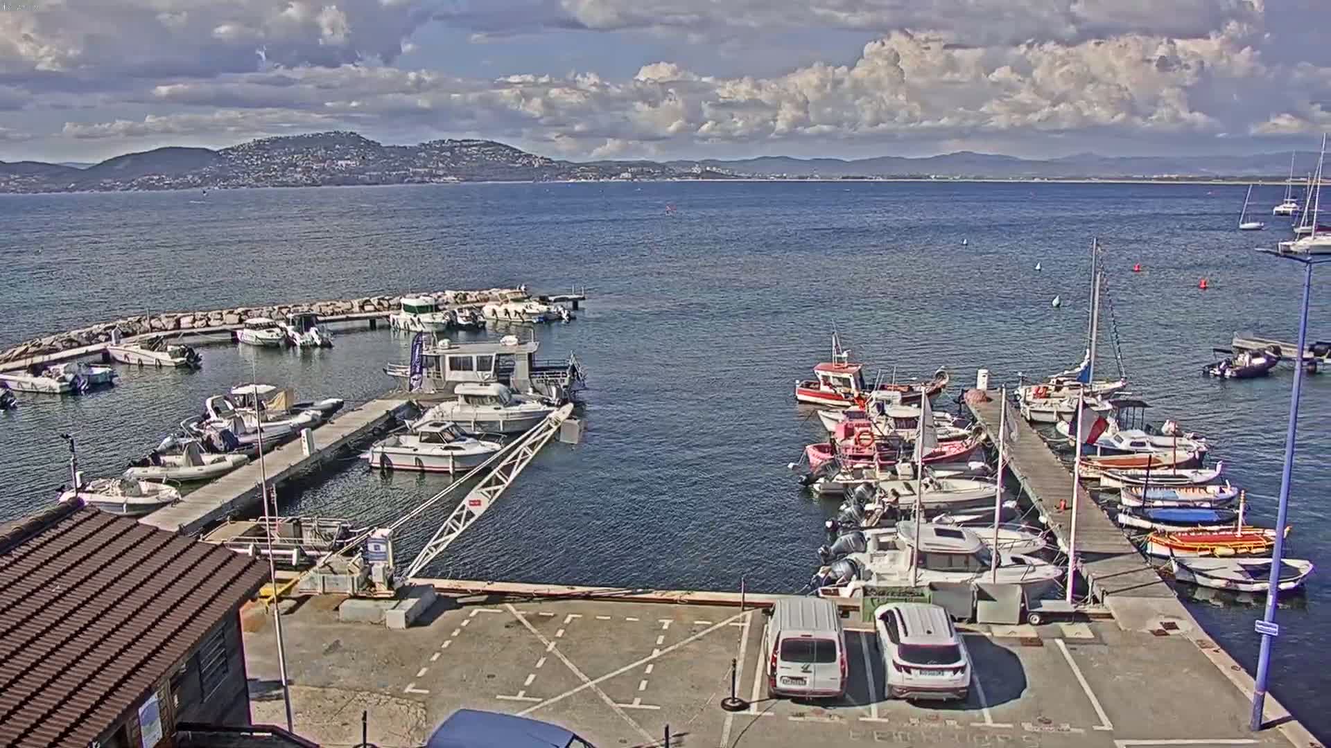 A marina filled with numerous boats of various sizes is situated beside a body of water under a partly cloudy sky, with a distant mountain range visible in the background.
