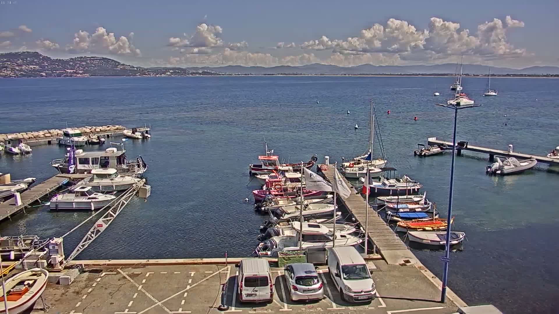 A marina filled with various boats and yachts is situated on calm, blue water under a partly sunny sky, with distant hills visible on the horizon.
